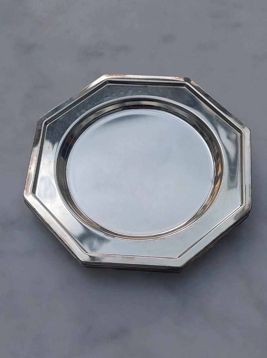 A Set Of 6 Coasters from Les Objoies, designed in a reflective silver octagonal shape with a smooth, shiny surface, is elegantly placed on a neutral gray background. These coasters, reminiscent of modern design and crafted from durable material, feature wide rims and slightly raised edges.