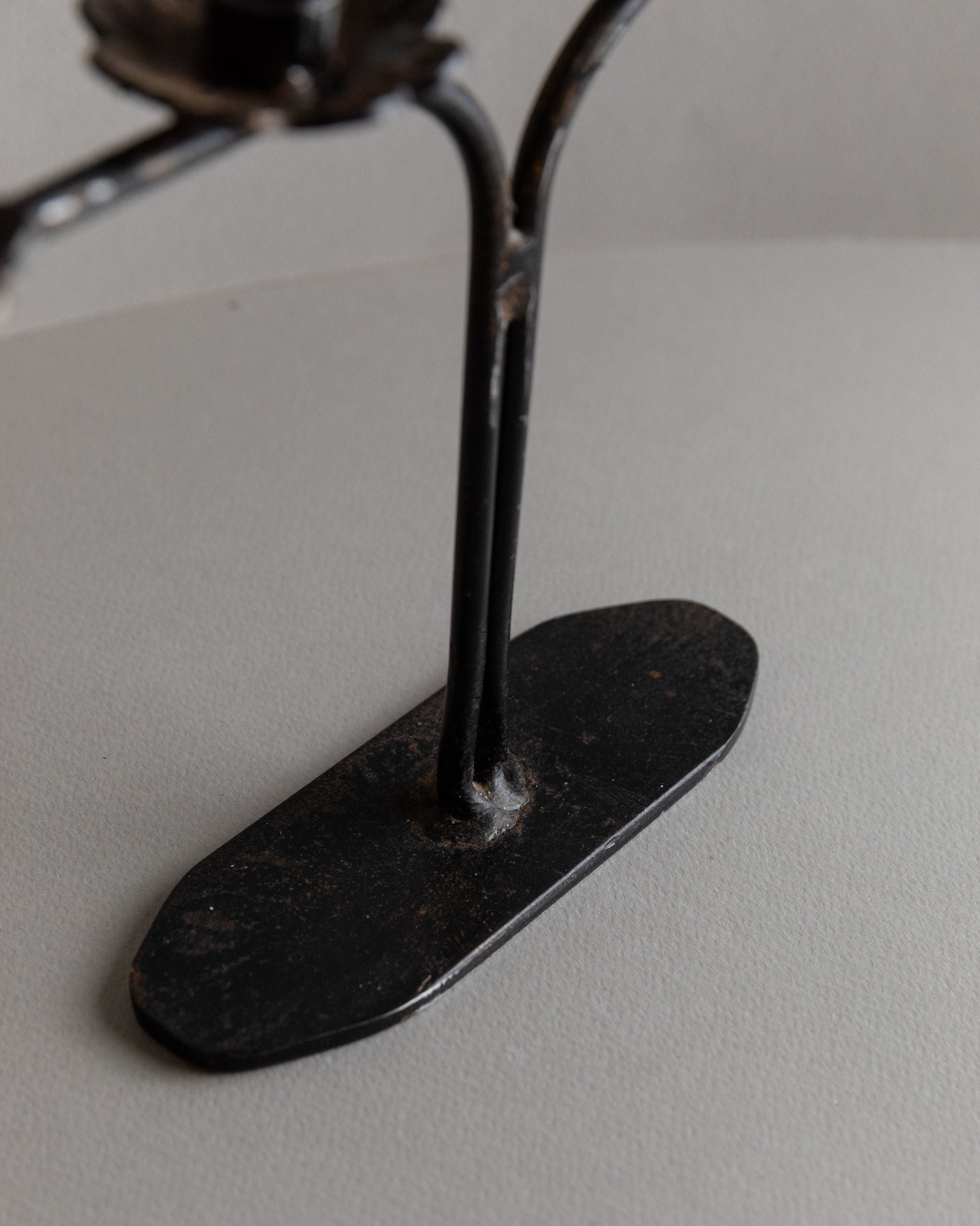 A close-up of the Bottega Jacobs Iron Candleholder, Italy 1960s, reveals its brutalist design with two vertical rods on an oval base. The black metal stand on a gray surface evokes a vintage iron candlestick, featuring a rugged and slightly textured surface for an intriguing touch.
