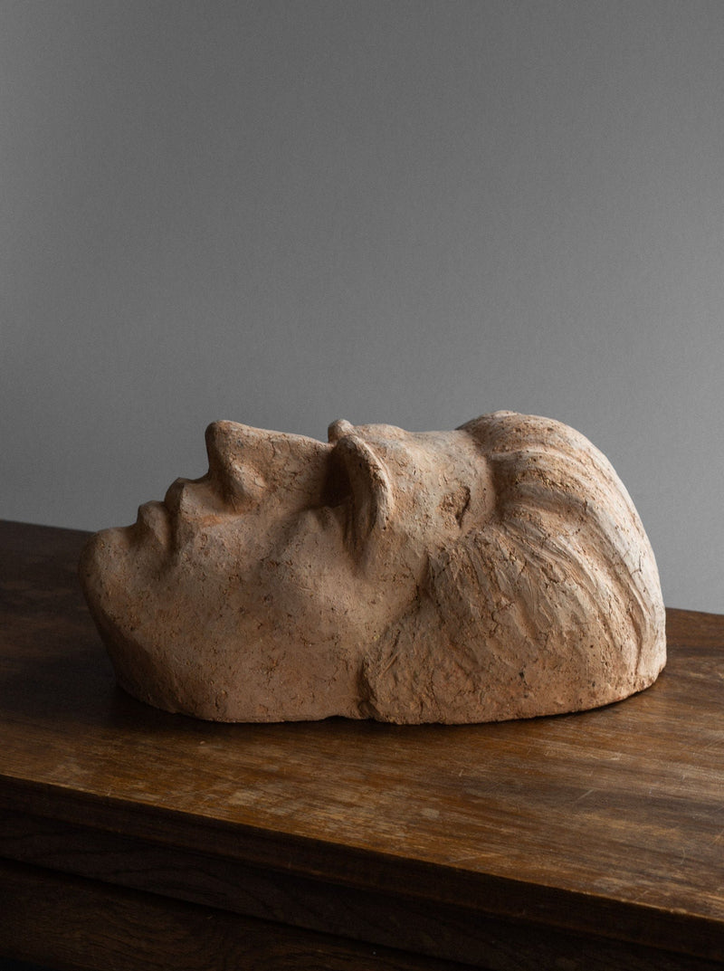 A vintage charm radiates from the Large Ceramic Portrait / Wall Sculpture, 1930s by Spigel, showcasing a serene human face lying on its side atop a wooden surface. The artwork, reminiscent of Holger Persson's style, captures a peaceful expression with closed eyes and finely detailed features including a textured hair pattern set against a plain, muted gray wall.
