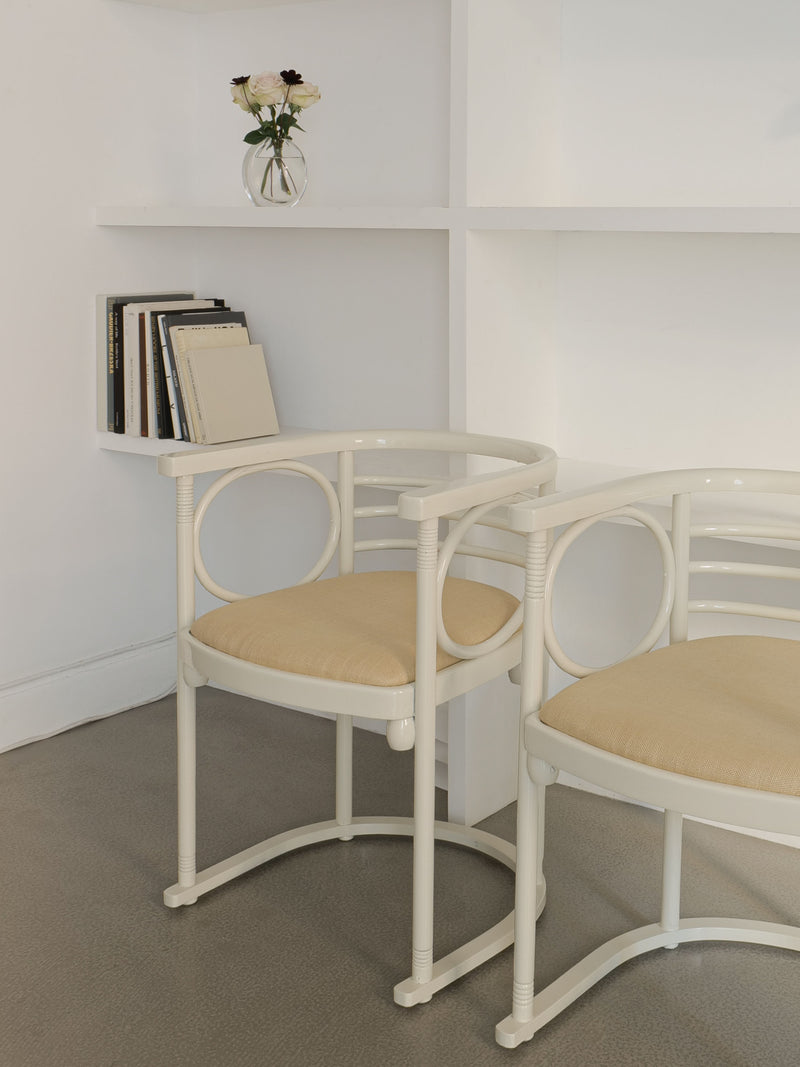 Two Thonet Bentwood Chairs by Josef Hoffman from Collection apart, featuring circular designs in a cream color, are placed against a white wall with built-in shelves. These lacquered bentwood armchairs enhance the minimalist and serene atmosphere, with the shelves displaying a small vase of flowers and several books.