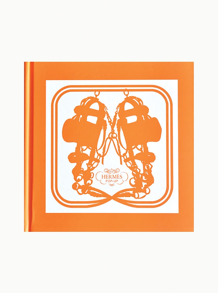 A Hermès Pop Up Book by Maison Plage with a white cover featuring an intricate design of intertwined ropes and harnesses in vibrant orange. The center showcases the text "Hermès Pop Up" in a decorative font, reminiscent of their luxury scarf designs. The symmetrical layout extends the striking orange around the edges.