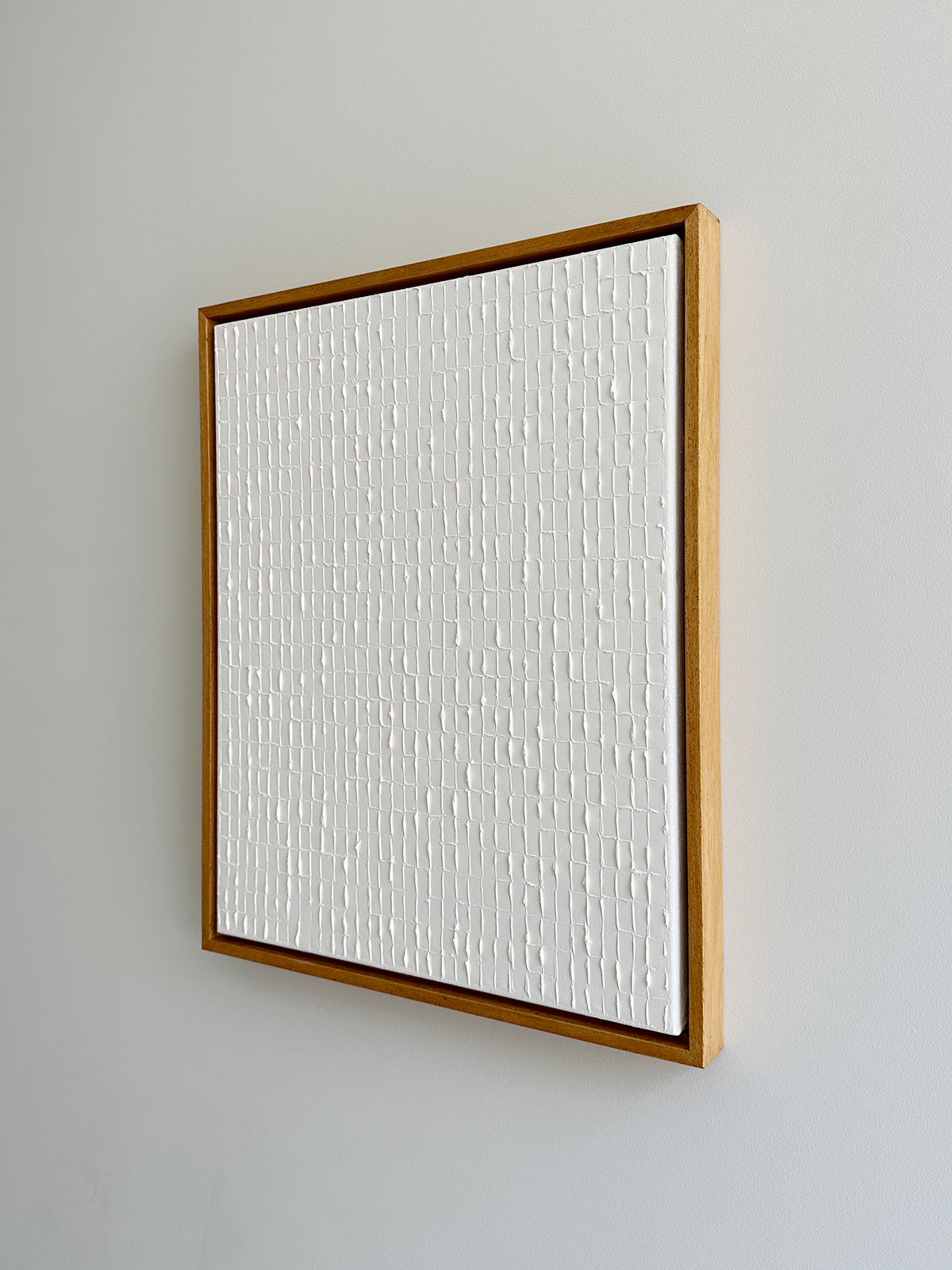 A textured white canvas framed in a simple wooden frame is mounted on a light gray wall. The canvas features a grid-like pattern created with raised, textured lines that add depth and dimension, reminiscent of the Korean Dansaekhwa movement's emphasis on texture and purity. This piece is titled "Warm White - Oil on canvas" by Roi Elmaliah.