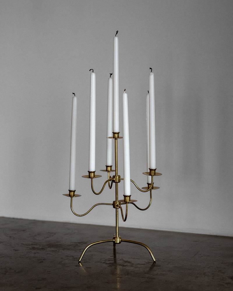 On a gray surface, Spigel's Swedish Mid-Century Brass Candelabra holds six tall white candles, exuding Scandinavian Modern elegance. The blurred background highlights this minimalist piece akin to a Josef Frank design.