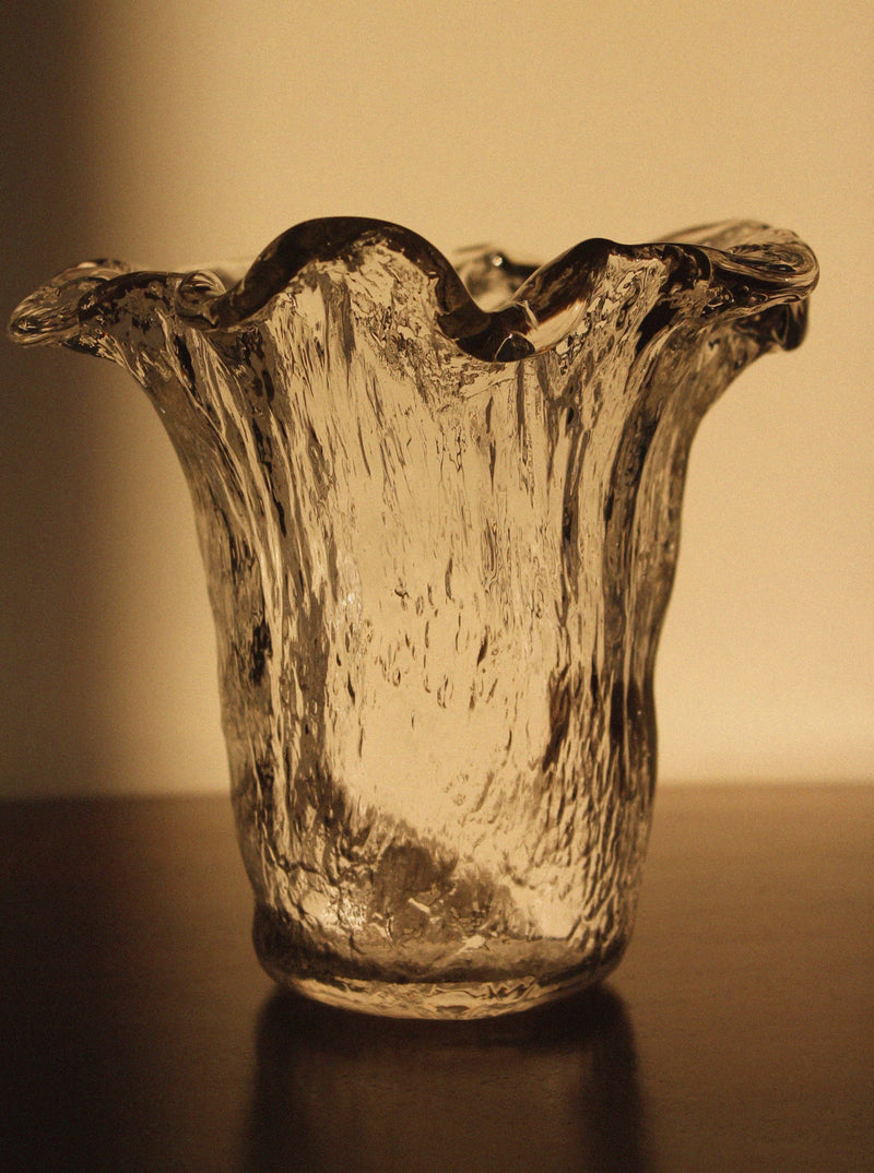 The Uno Westerberg 60s Organic Glass Vase by Vintage Stories sits on wood, its textured, ruffled rim aglow in warm light that highlights its vintage design and casts elegant shadows.