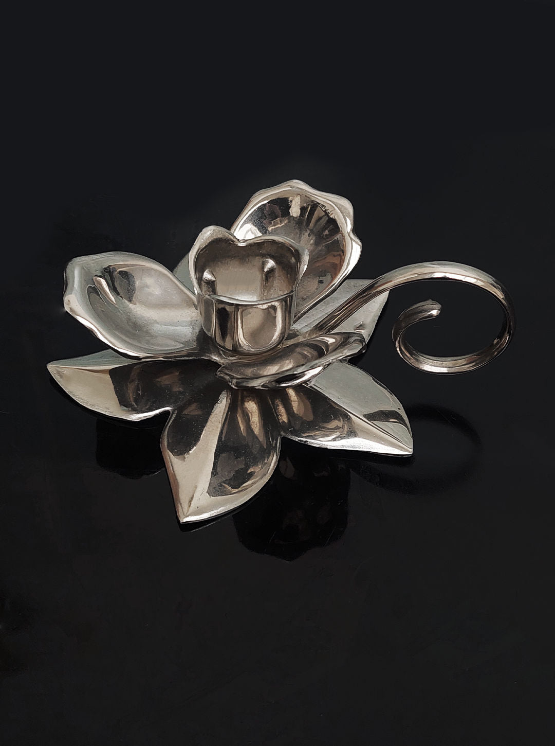 Inspired by nature, the Floral Candleholder from Les Objoies features silver-plated craftsmanship resembling a blooming flower with a gracefully curved handle, all set against a dark backdrop.