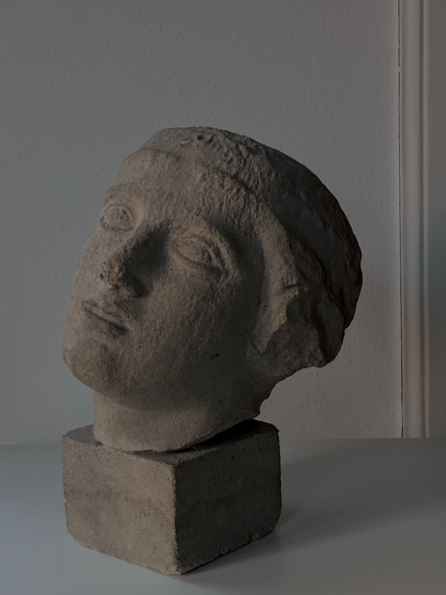 Displayed on a straightforward square base, the Decorative Bust by Médecine showcases mid-20th-century artistry. Its classical features in sandstone composite reveal a calm expression with a subtle tilt, against a plain light gray wall. The soft, natural lighting highlights the piece's elegance as a decorative sculpture.