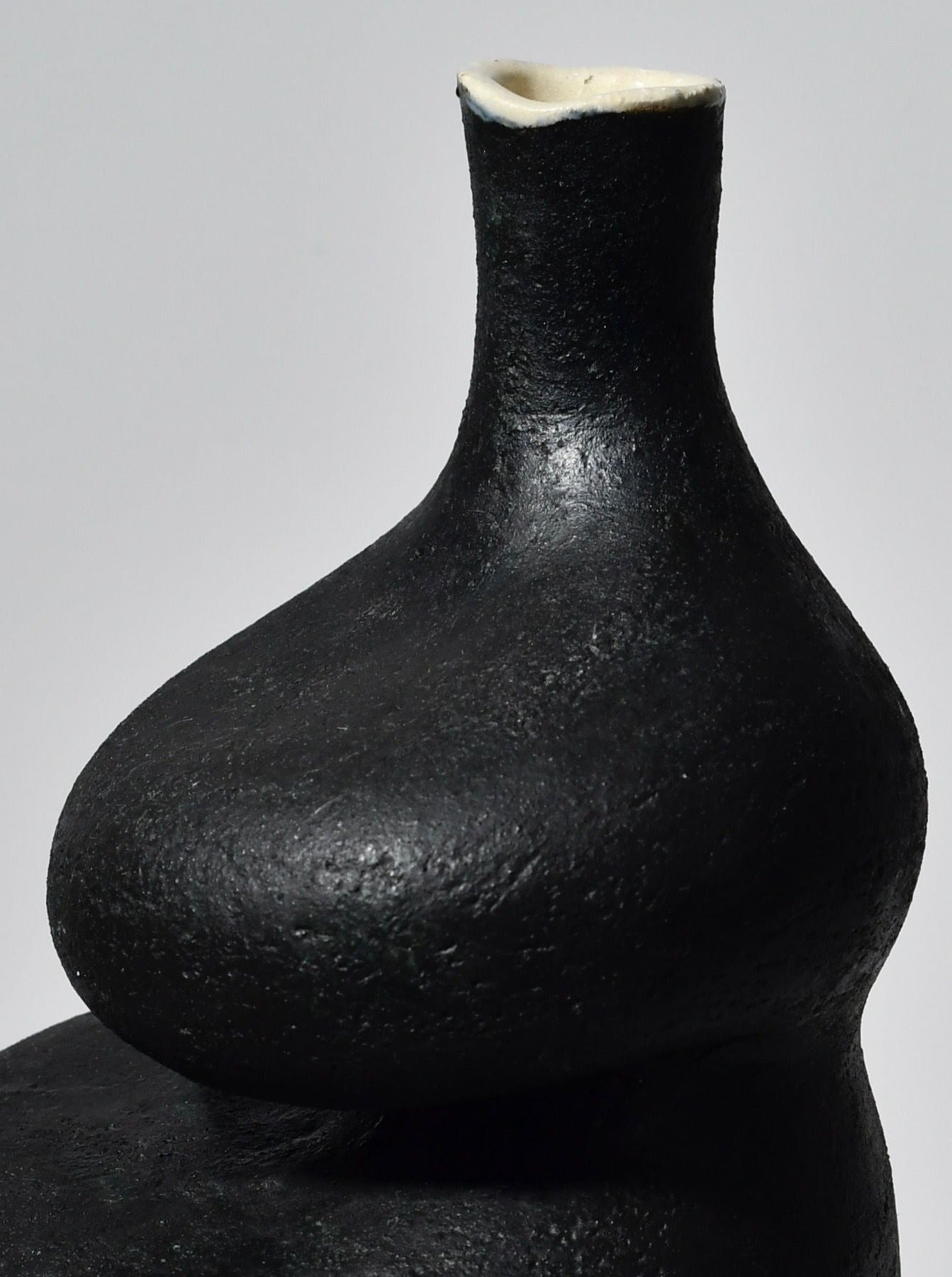 Close-up view of a sculptural vessel with a textured black surface and smooth, narrow neck, showcasing Lucia Mondadori's Black Lilith with Oxide Drip's subtle shine and simplistic, elegant shape.