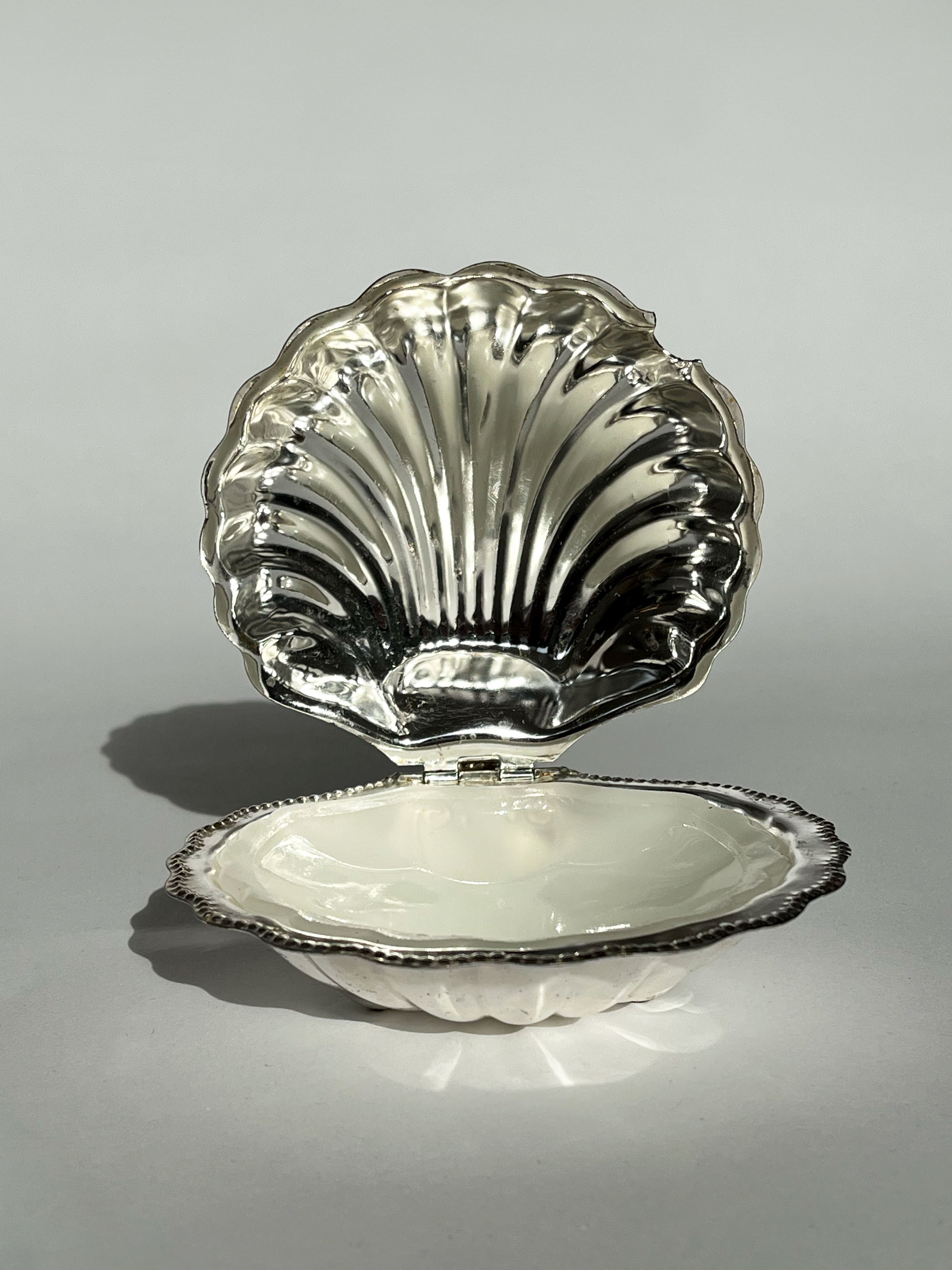 A decorative Shell Shaped Butter Dish by septembre studios, featuring a pearly white interior and polished silver exterior, is displayed against a neutral background. The reflective surface highlights its elegant design, reminiscent of vintage brass butter dishes.