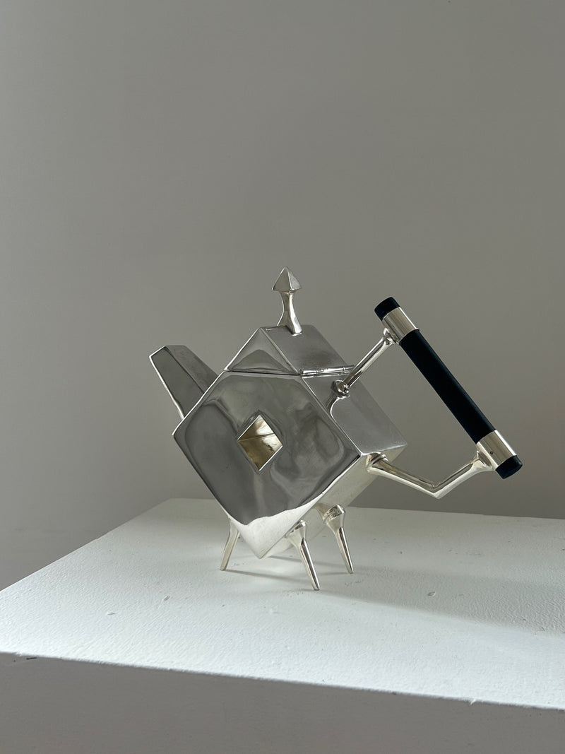 The Christopher Dresser Square Teapot by Porter Studio, a sleek silver-plated piece with geometric shapes and sharp angles, features a black handle and is elegantly displayed on a plain white pedestal against a light gray background.