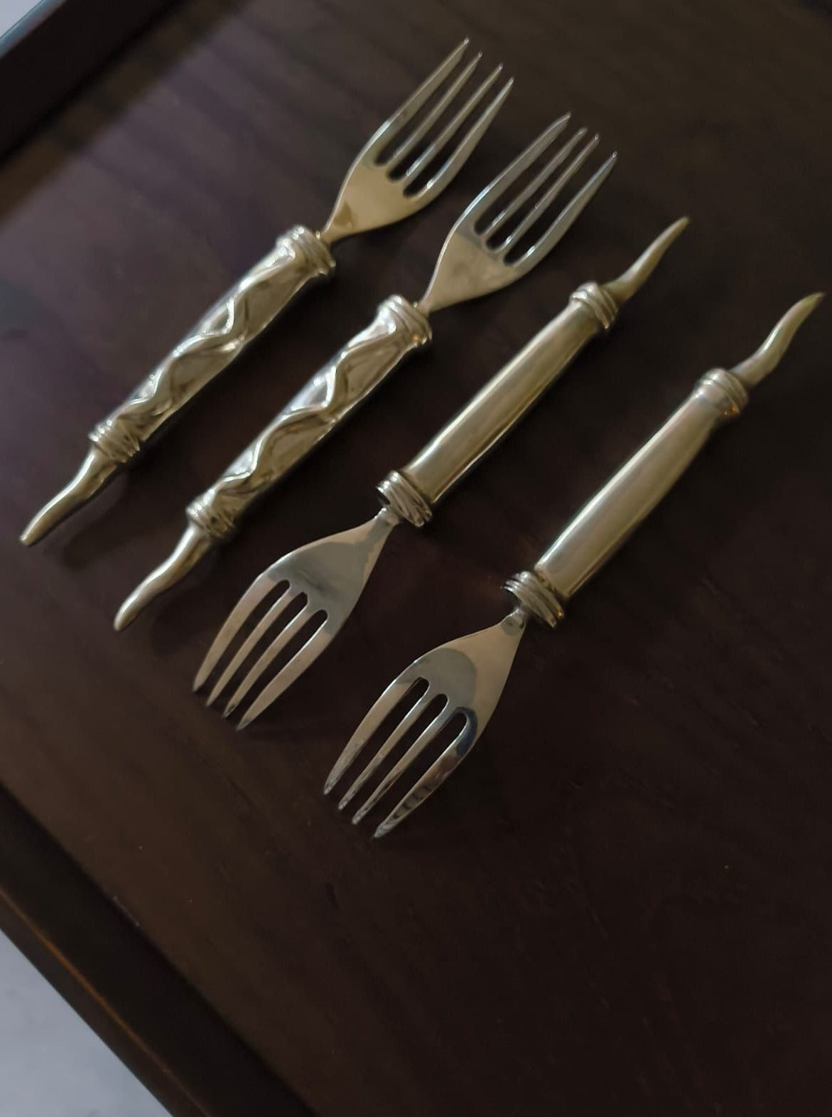 Four antique-style forks with ornate, twisted handles and two-pronged tines are placed on a dark wooden surface. The Rare Vintage Cutlery Set by Médecine includes a set of 4 forks and 4 stainless steel knife blades, all featuring decorative designs that lend an elegant appearance.