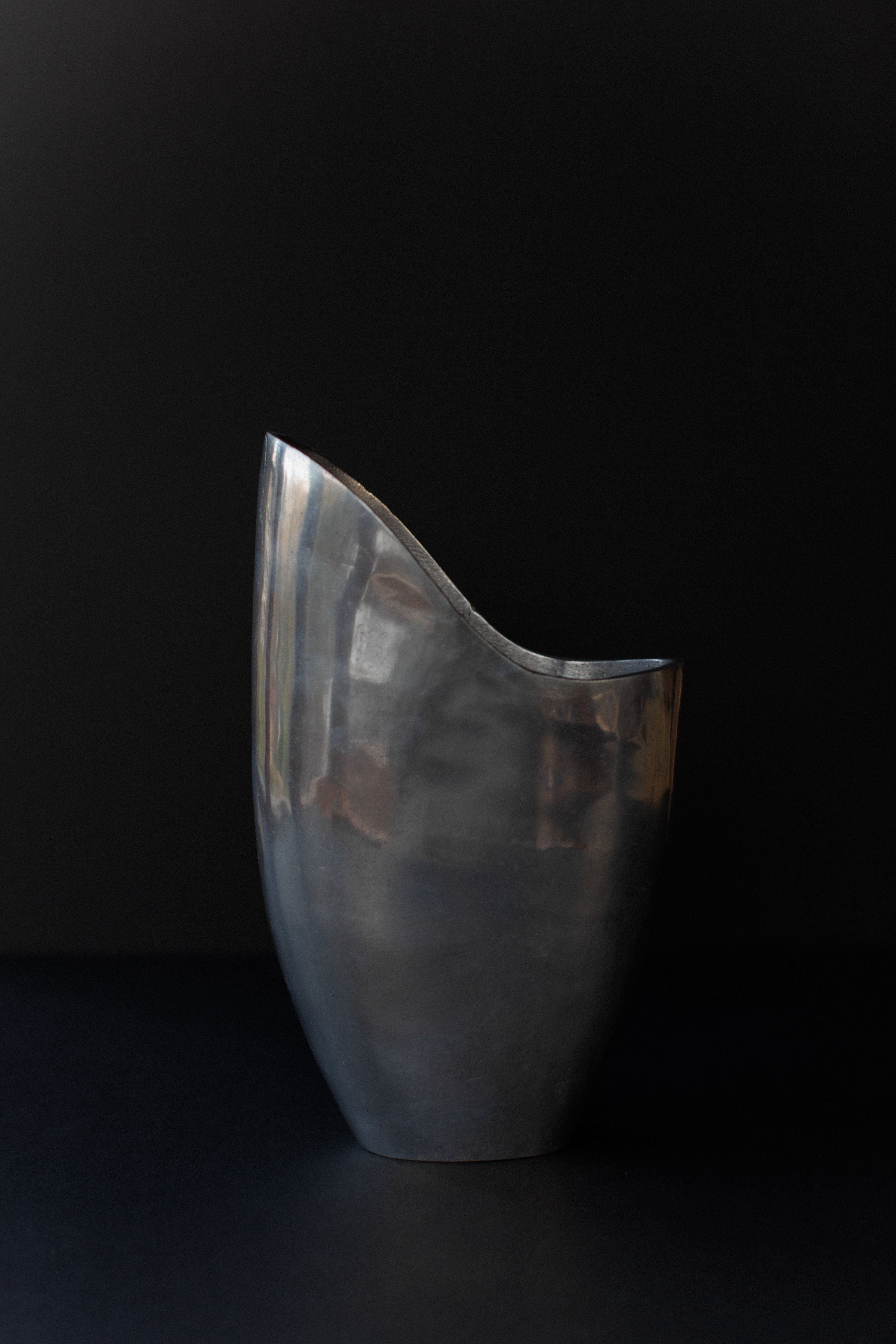 The Out For Lunch Aluminum Cast Vase, with its sleek silver design and smooth finish, effortlessly complements a dark background. Its curvaceous form and asymmetrical opening evoke a modern industrial aesthetic and organic elegance.