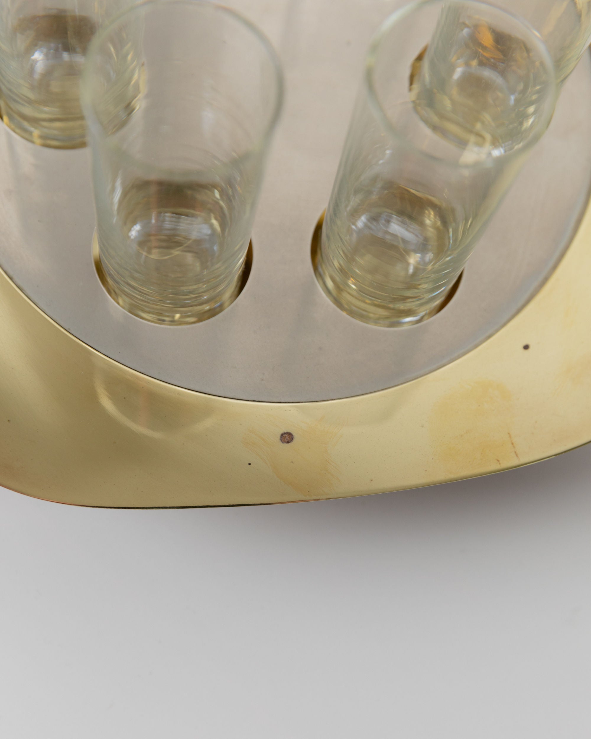 A close-up of a Bottega Jacobs Set Shot Glasses and Tray - Mid Century Design 70s shows a circular metallic tray with five slots, each holding a vintage shot glass. The tray has a golden finish with visible tarnish and dark spots. It rests on a light grey surface.