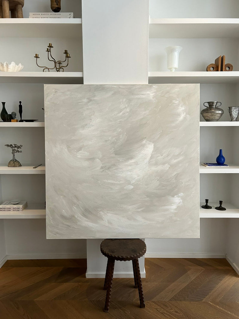 A large "Waves of Silence" painting by Mattias Storm, featuring swirling gray and white patterns, rests on a small round wooden stool against a white wall. Crafted on canvas, this masterpiece is accompanied by shelves in the background adorned with vases, books, and a candle holder.