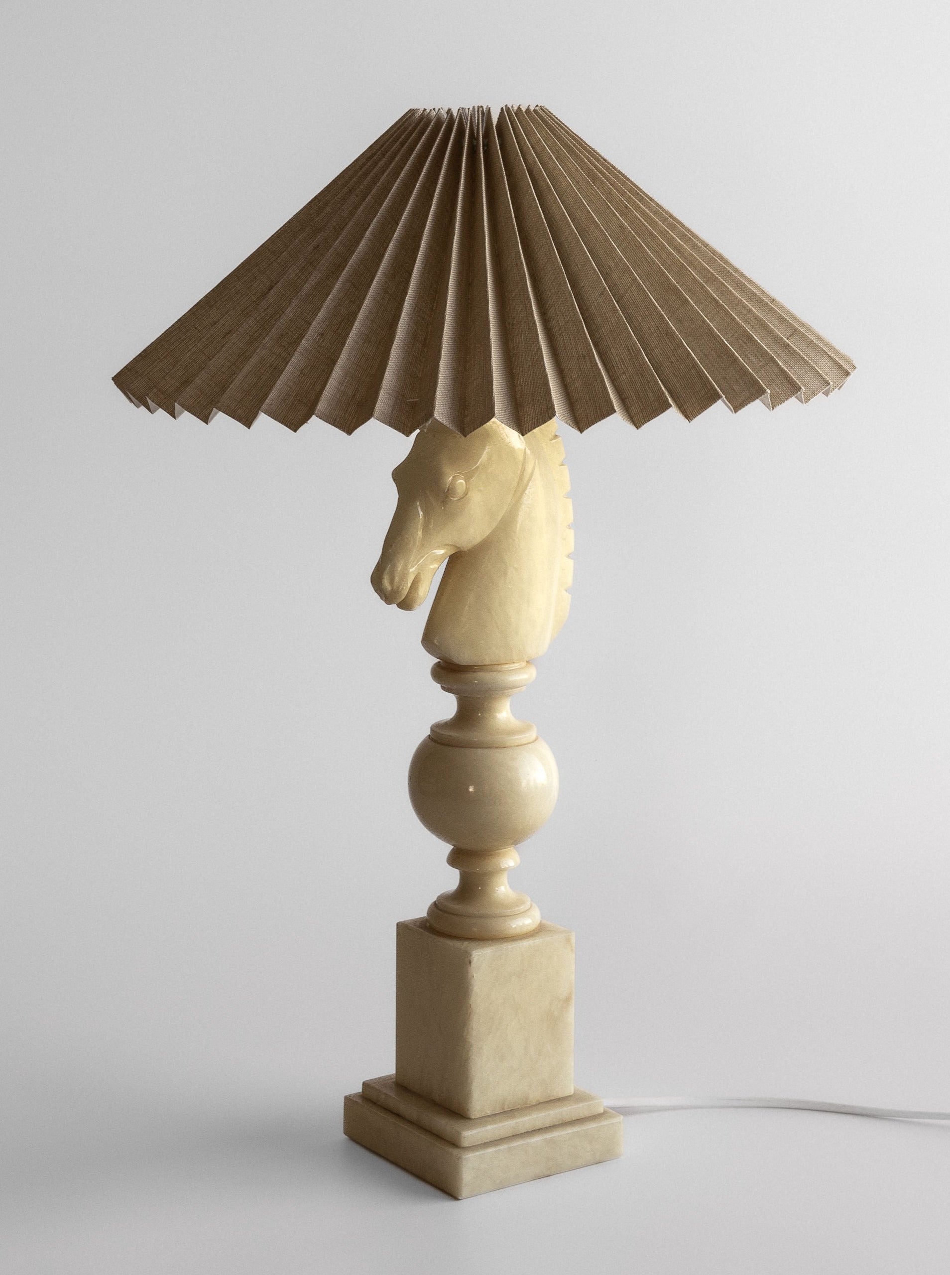 An ivory-toned alabaster lamp from Spigel, dubbed the "White Knight Alabaster Table Lamp," originating from 1970s Italy, features a pleated beige lampshade. The handcrafted base showcases a sculpted horse head atop a pedestal, imparting a unique and artistic flair that evokes vintage Italian lighting. The plugged-in lamp rests on a white surface against a neutral background.