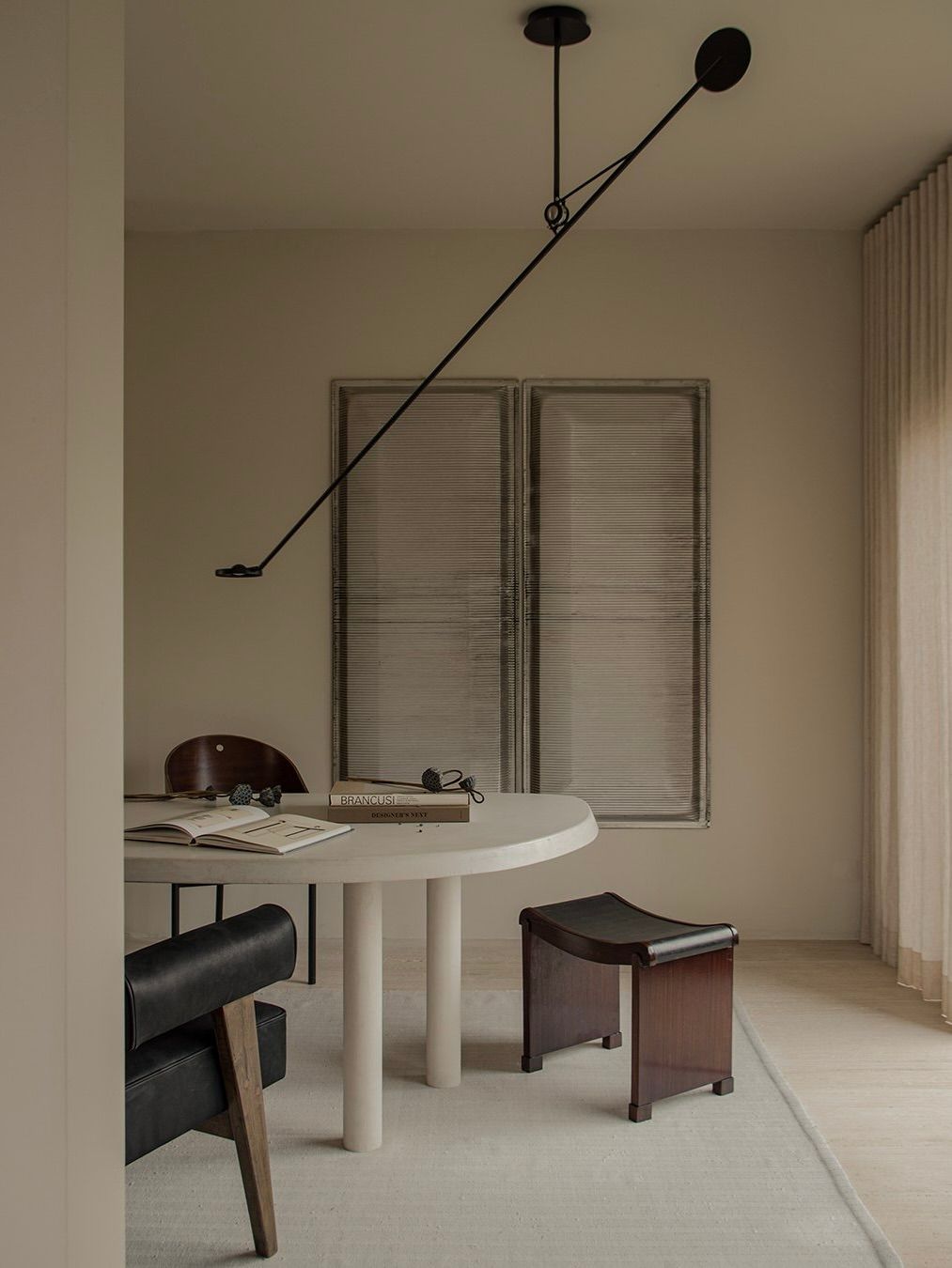 A minimalist room features a round white table with a modern black lamp. A black chair and Bicci de' Medici's Pierre Chareau Curule (Model MT 1015) Stool surround the table. Books and headphones rest atop it, while two abstract Art Deco paintings adorn the beige wall beneath sheer window curtains.