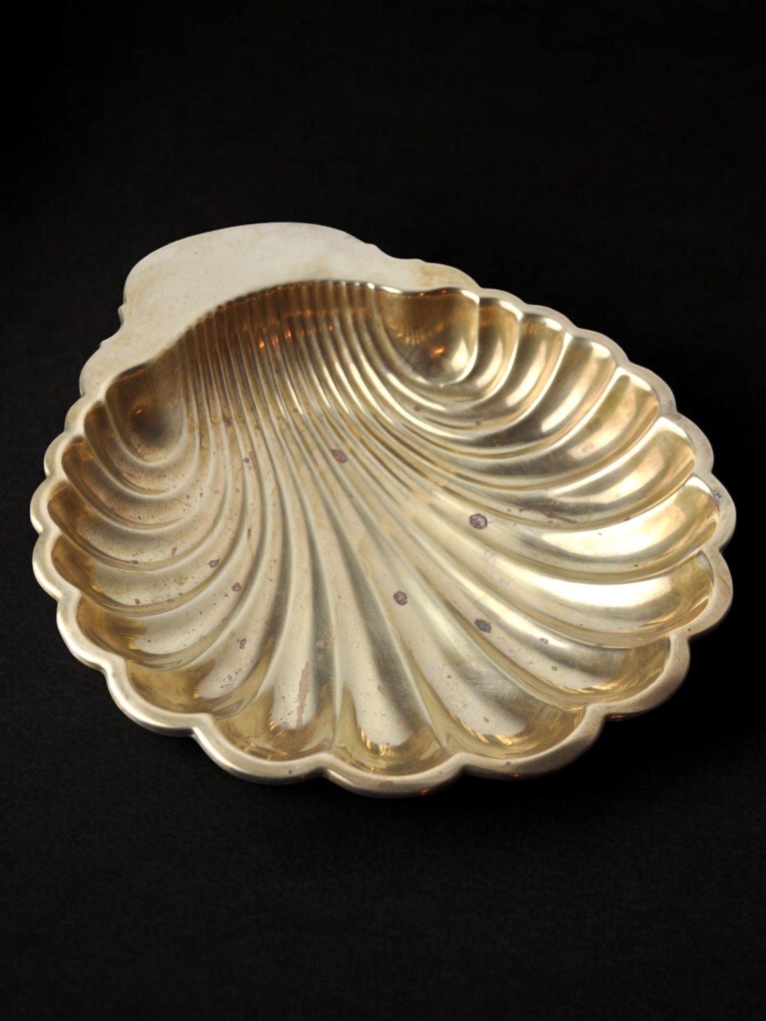 The Large Shell Shaped Vide-Poche 70s by Malamar Studio features a gold-colored, shell-shaped dish adorned with ridged patterns and a scalloped edge, set against a black background. Made with a brass structure and designed in Italy, the surface displays tarnish spots that contribute to its vintage charm.