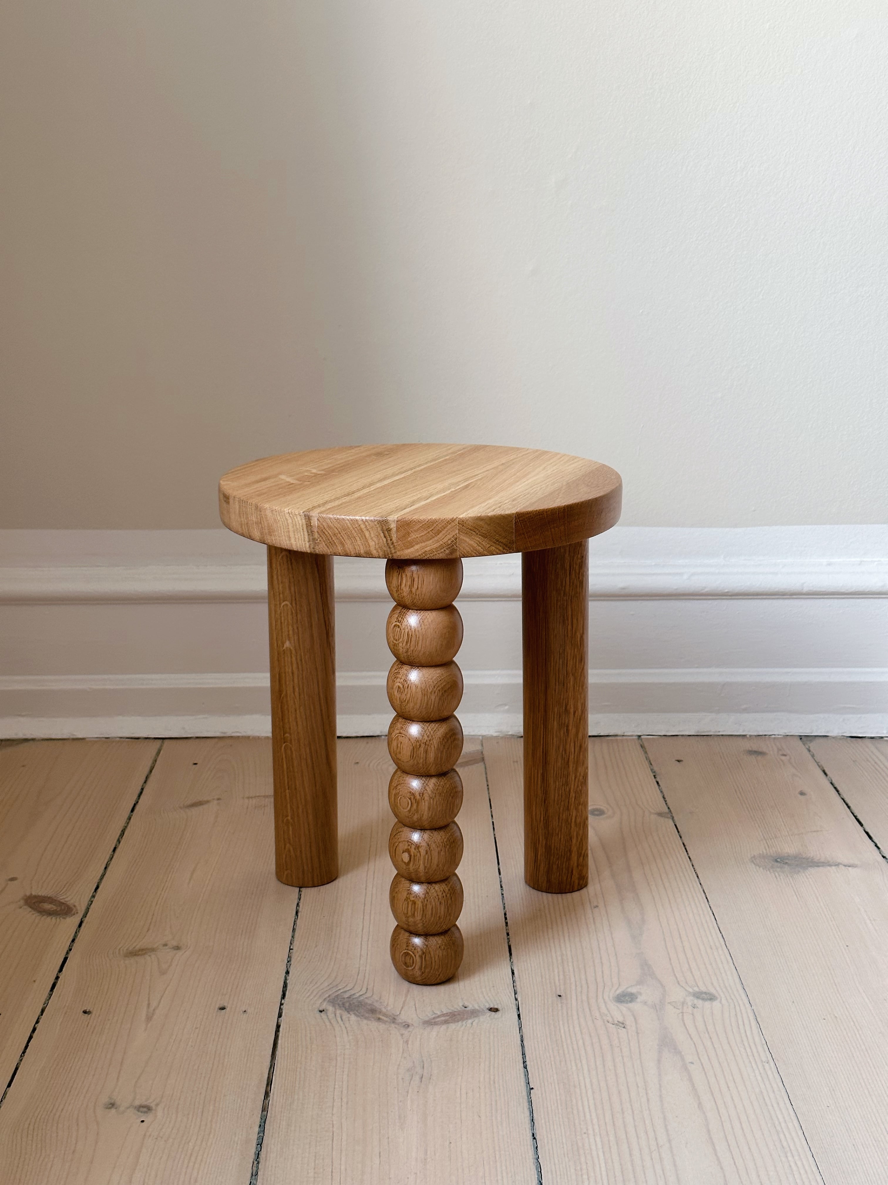 The Piloti Side Table in Oak by Maitoinen Home, a small round wooden stool, stands on a light wooden floor. Two legs are simple and cylindrical, while the third features Euclidean geometry with stacked spheres. The backdrop is a plain light-colored wall.