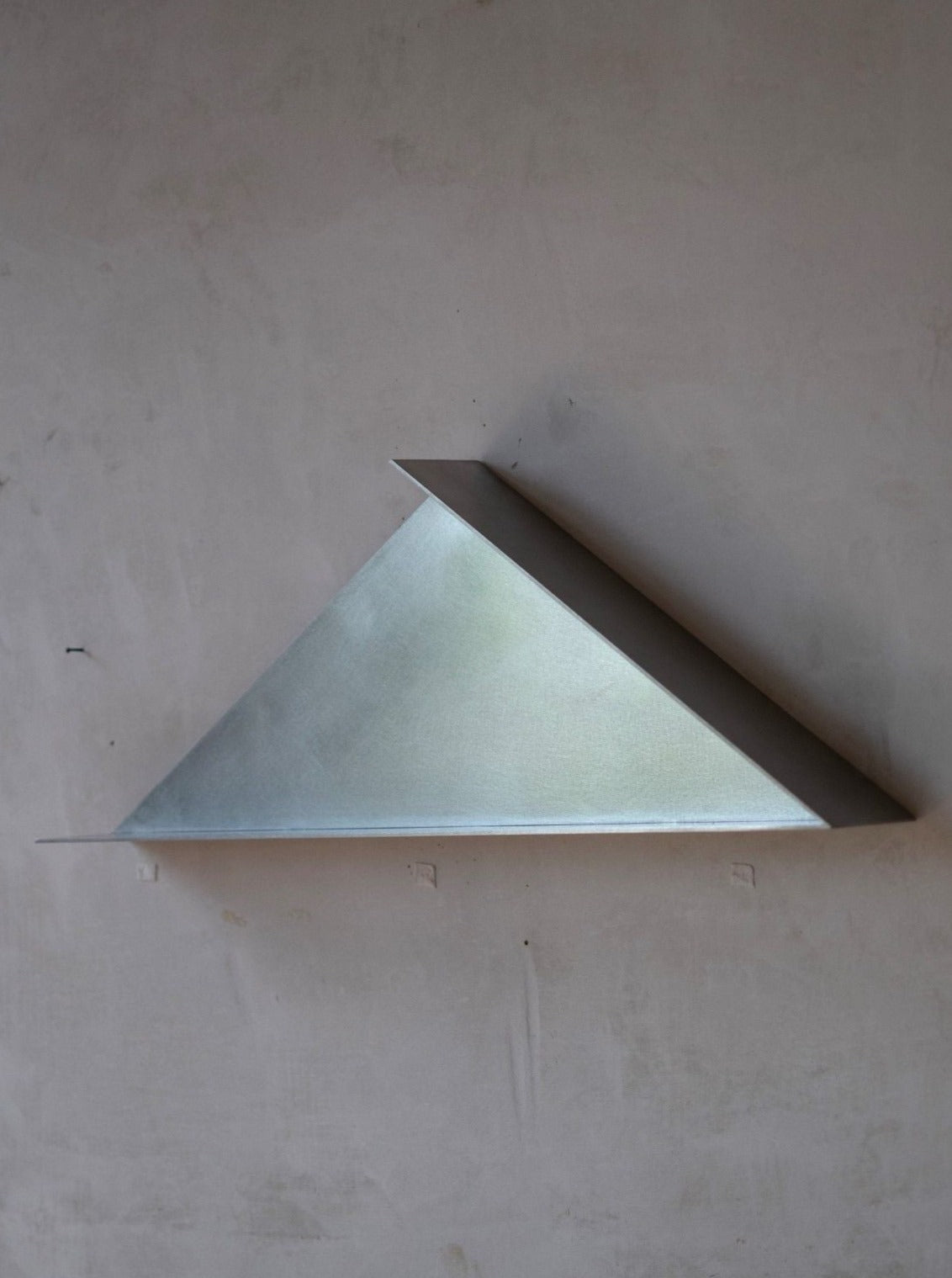 A minimalistic, triangular stainless steel wall shelf is mounted on a grey wall. The shelf has a simple, angular design, with the right-angled triangle creating a modern, sleek appearance. The smooth metal finish adds to the contemporary aesthetic of this limited edition architectural decorative element: *The Front - Shelf* by **COSEINCORSO**.