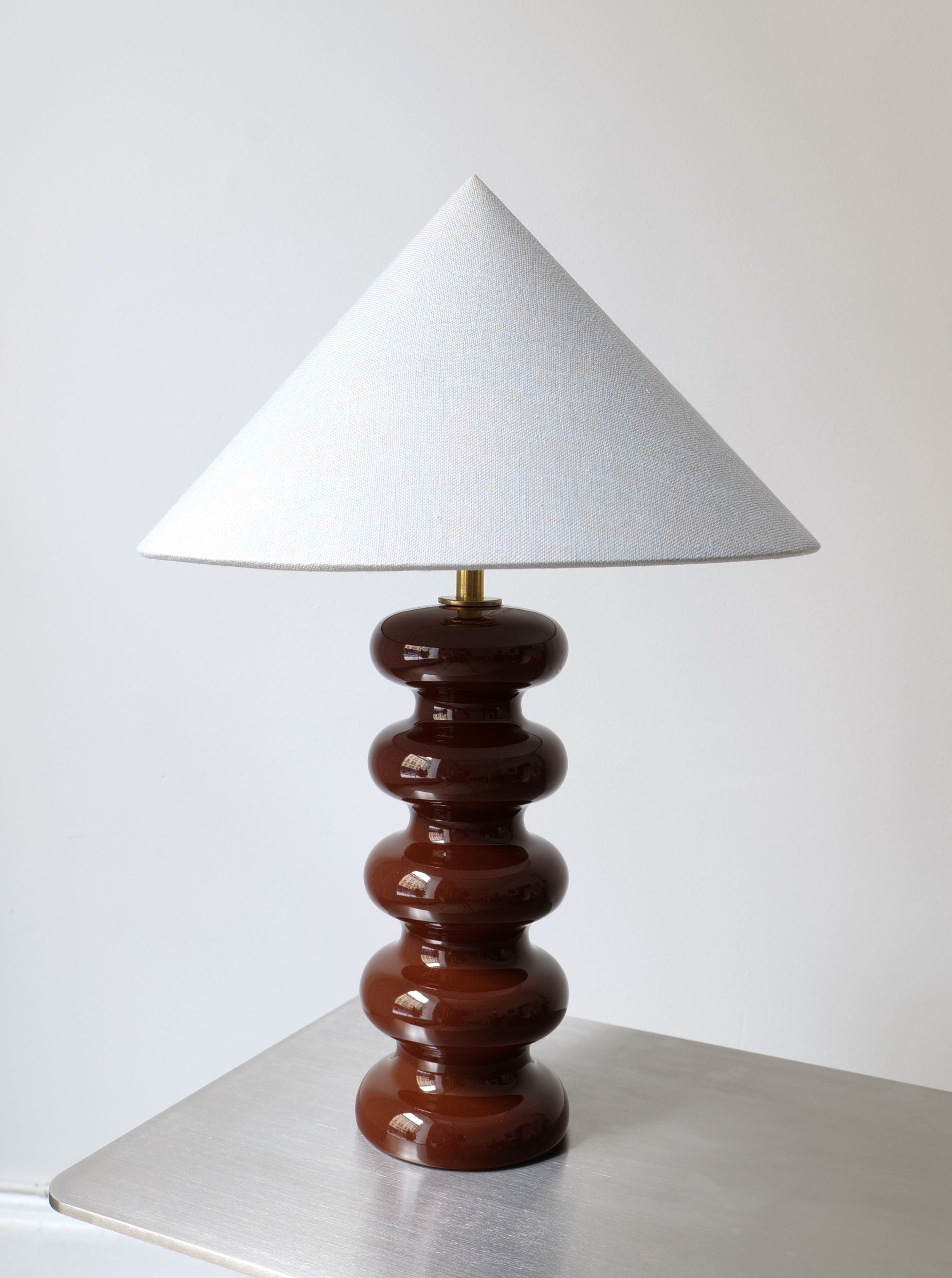 A contemporary table lamp featuring a glossy, segmented, brown ceramic base and a handmade cone-shaped white fabric lampshade. The Sculptural Ceramic Lamp by Collection apart is placed on a simple, flat, metallic surface against a plain white wall, creating a minimalist aesthetic.