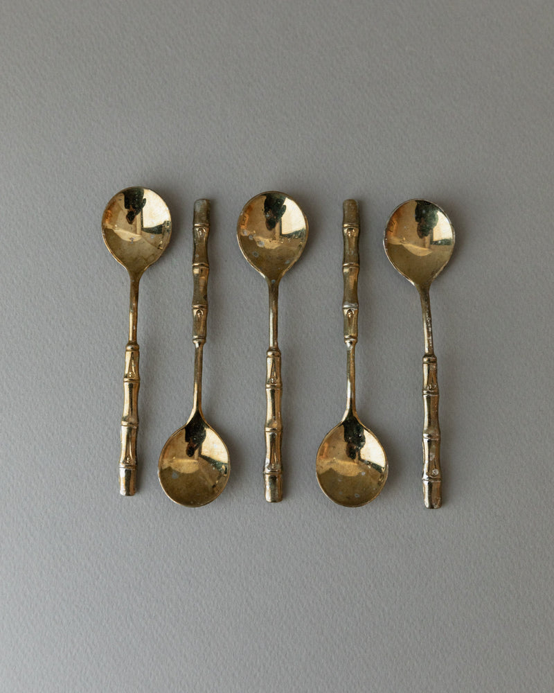 A row of five gold-plated teaspoons by Bottega Jacobs from the 1970s, featuring textured handles on a light gray surface, captures the glamour of Hollywood Regency style.