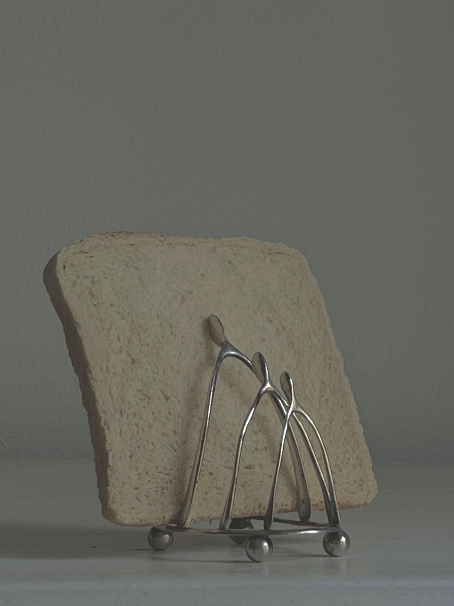 A slice of bread rests in a Wishbone Silver Toastrack by Médecine, featuring curved bars on a plain surface against a muted gray background.