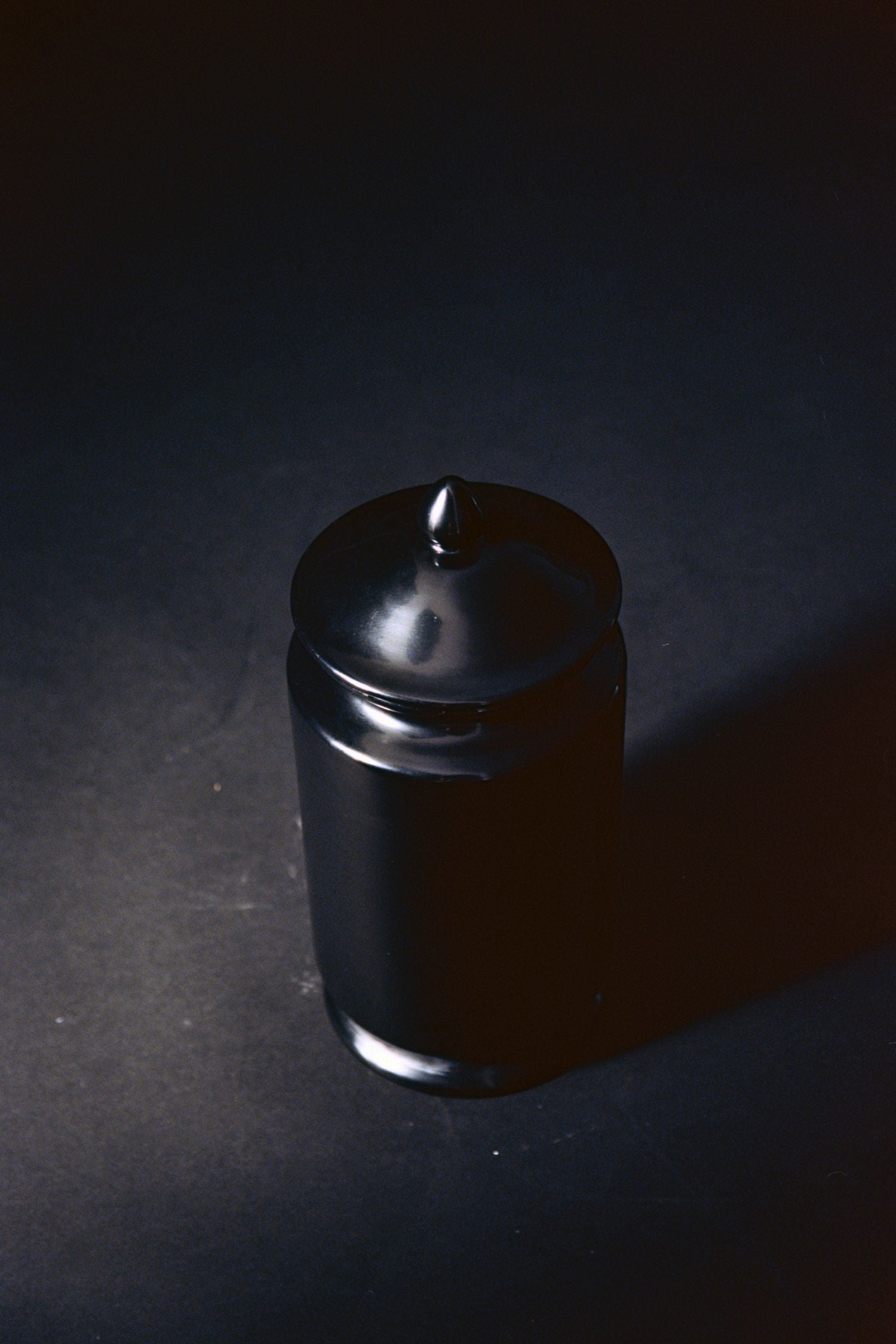 The "Albarello" Jar by COSEINCORSO, a glossy black ceramic apothecary jar with a lid, rests on a dark surface, its smooth finish softly illuminated and reflecting gentle light.