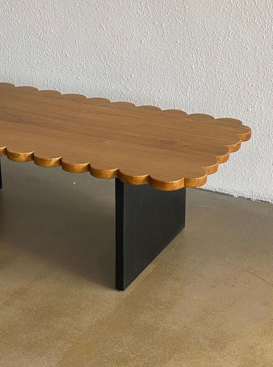 The Biskut Coffee Table by Studio Kallang is a modern solid teak coffee table with unique scallop detailing on its surface. The handcrafted table boasts a light wood finish on top and black V-shaped legs, placed against a light-colored wall on a concrete floor.