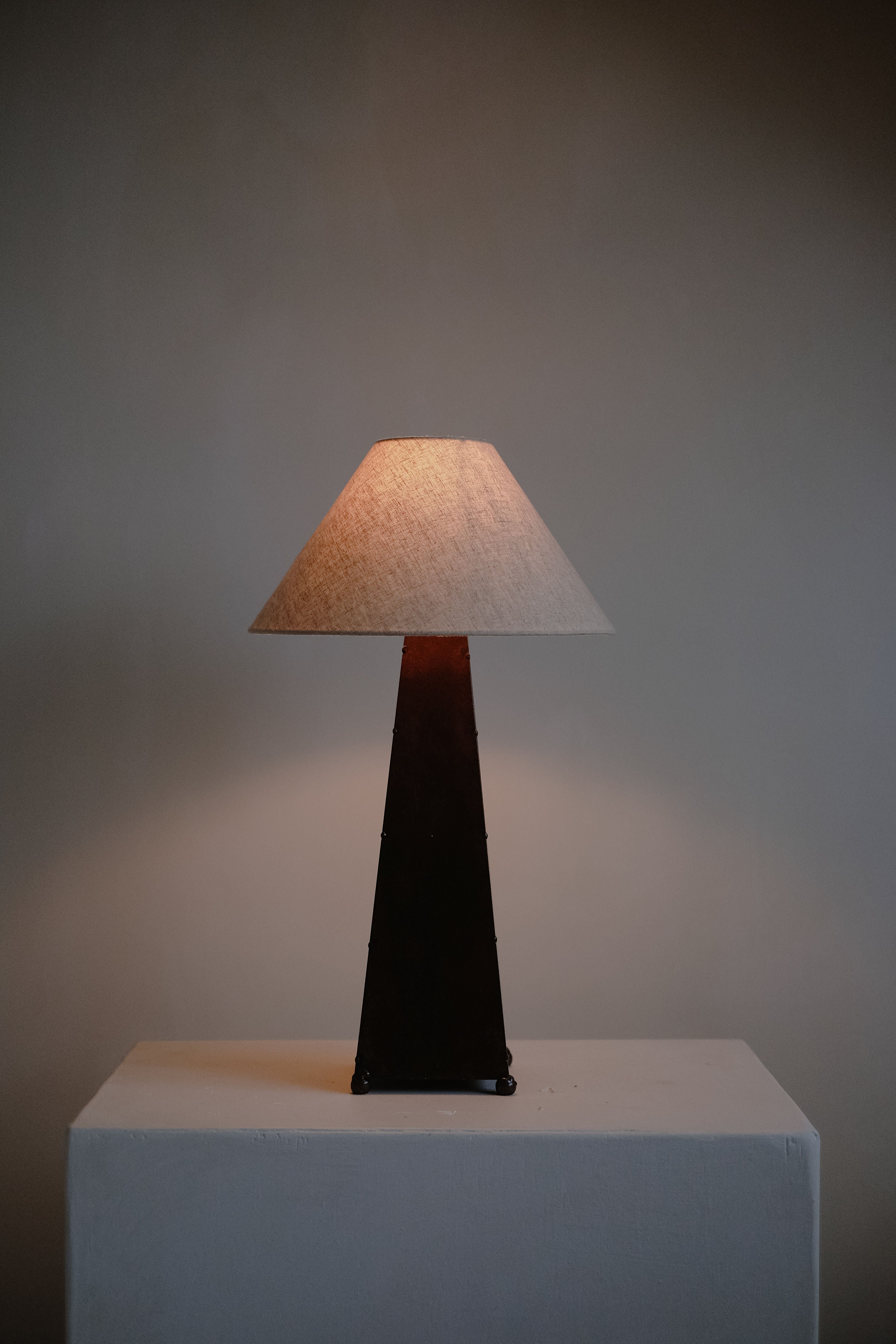 The Obelisk Table Lamp by septembre studios, with its rust-copper patina and dimmable switch, elegantly stands on a triangular base and conical shade, casting a warm glow. It's placed on a simple light-colored rectangular surface against a plain wall.