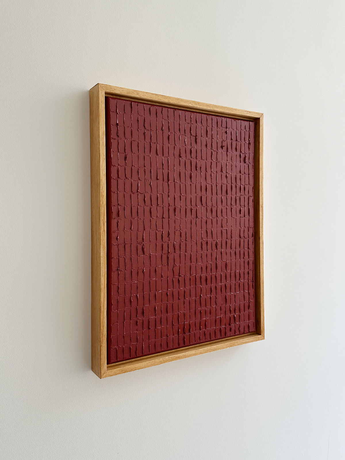 A wooden framed artwork is displayed on a white wall. The piece, inspired by Korean Dansaekhwa, consists of a textured, brick-like pattern in dark red. The rectangular frame is simple and adds a rustic touch to the abstract composition, inviting mindfulness through its organic forms. This exquisite work is titled "Indian Red - Oil on canvas" by Roi Elmaliah.