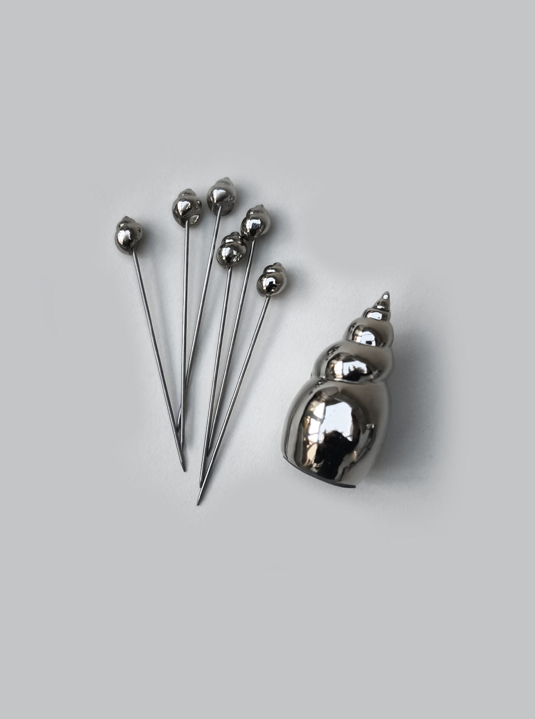 Six Les Objoies Shell Cocktail Picks with silver ball heads are displayed beside a shiny seashell-shaped holder, creating an ocean-inspired scene on a light gray background.