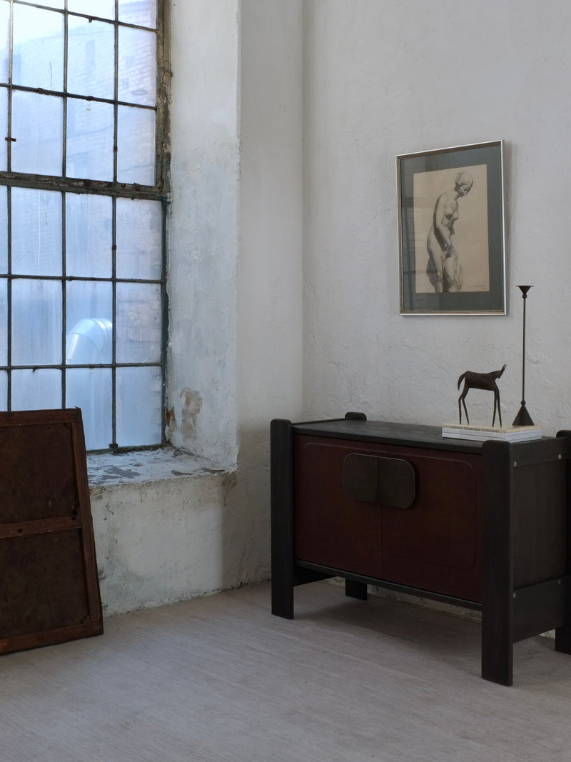 A minimalist room features a large industrial-style window, a small black cabinet with a horse statue, and a tall candleholder. An antique "Ebba Von Wachenfeldt Charcoal Drawing 1883" by Veter Vintage hangs above the cabinet, enhancing the room's weathered artistic charm.