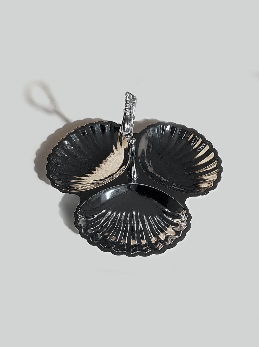 The Triple Shell Server by Les Objoies features a decorative design with three black, stainless steel sections that resemble sea shells and have reflective surfaces. A shimmering silver handle adorns the center, while the dish is set against a plain gray background.