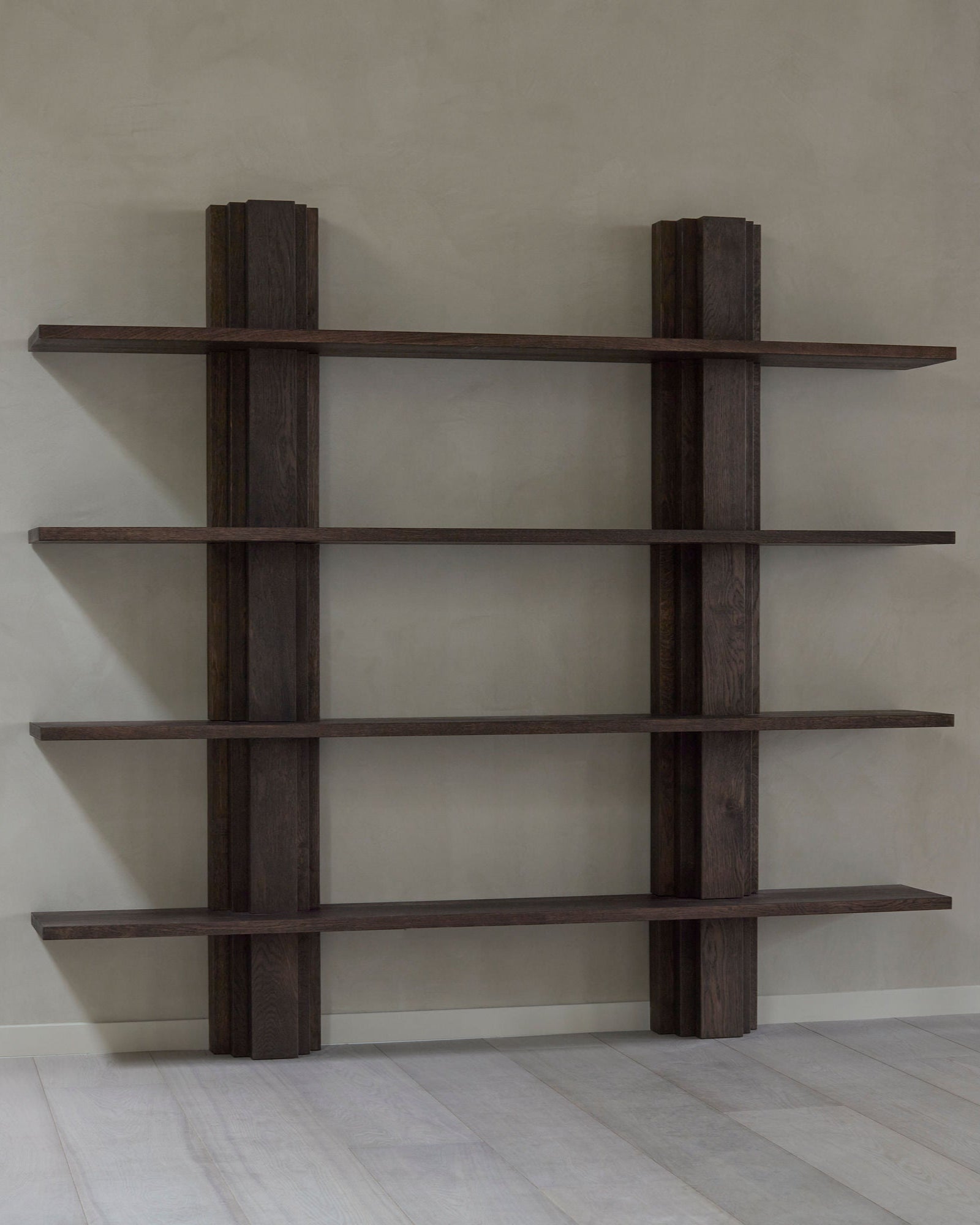 The Brut Bibliothèque by Mokko is a dark wooden bookcase crafted from solid European oak, featuring a minimalist design with four shelves and three vertical beams, part of the Brut collection, set against a light gray wall on a light-colored floor.