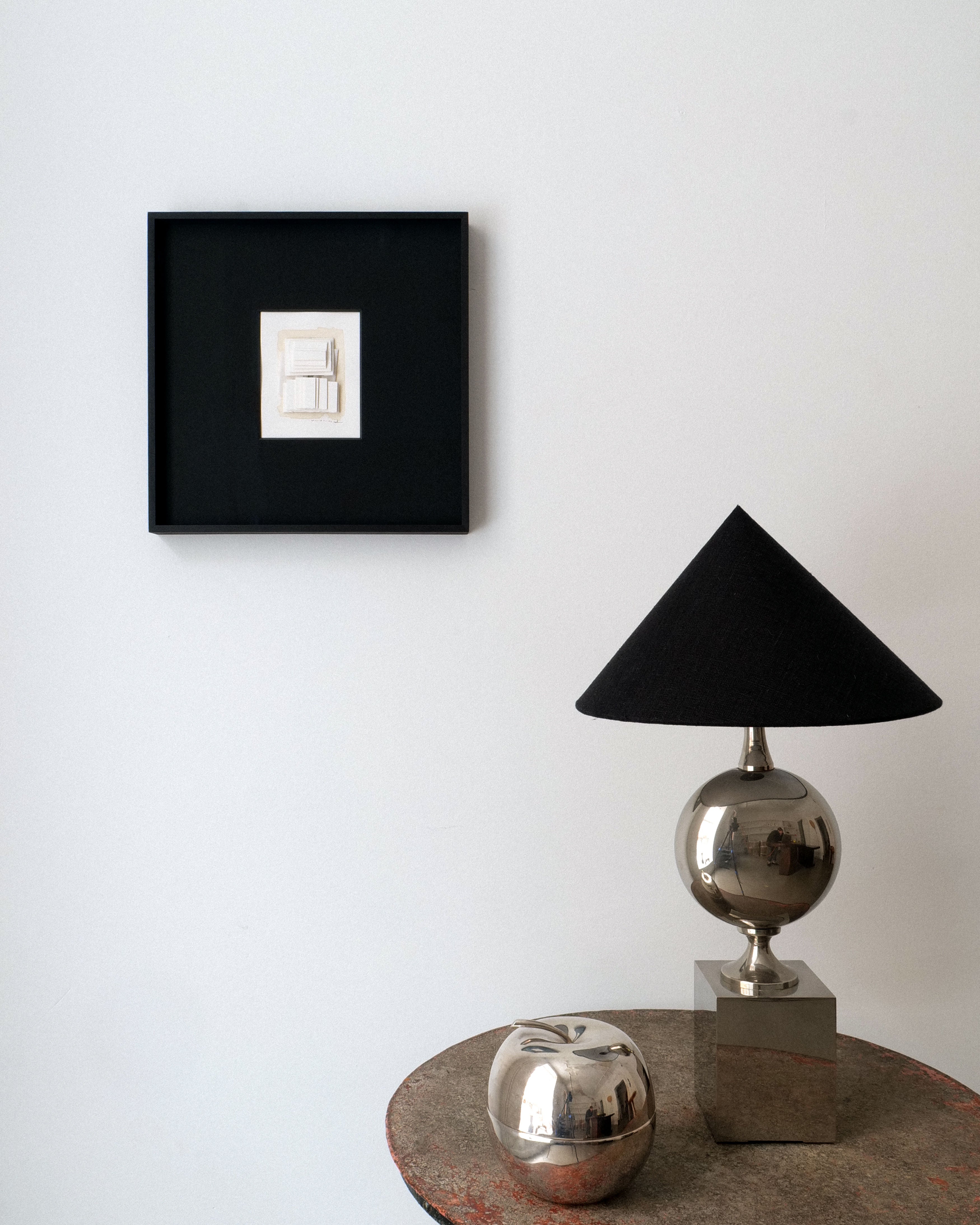 A minimalist interior features the Collage No.03 by Rasmus Wingårdh from Collection apart on a white wall. Below, a small round table displays a metallic silver lamp with a black shade and a reflective silver apple-shaped ornament.