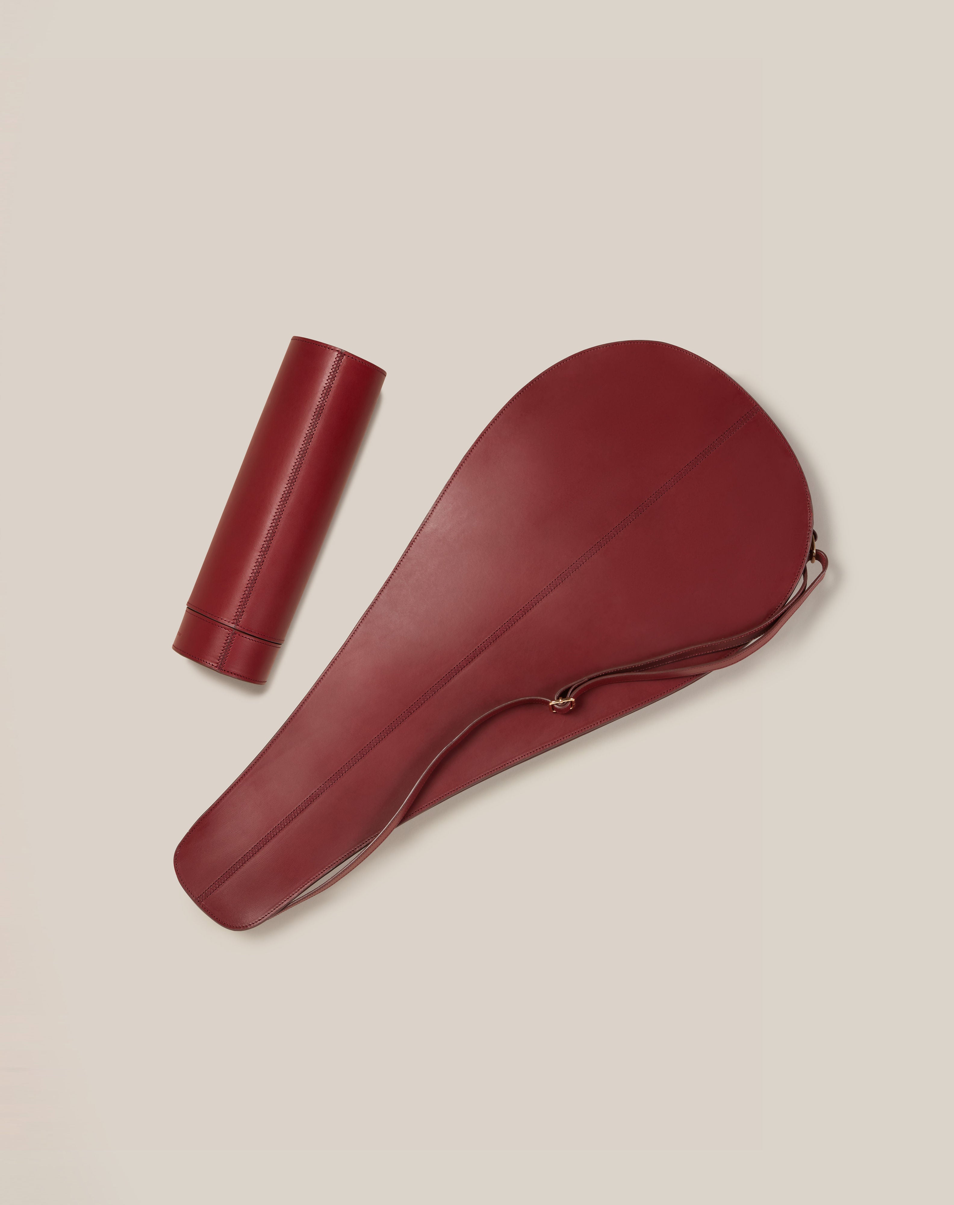 A maroon leather saddle cover made from sustainable vegetable-tanned leather and a matching Paradise Row Leather Tennis Racket Cover sit side by side on a light beige background. The bespoke British design features a handle and stitching detail, while the racket cover remains elegantly plain.