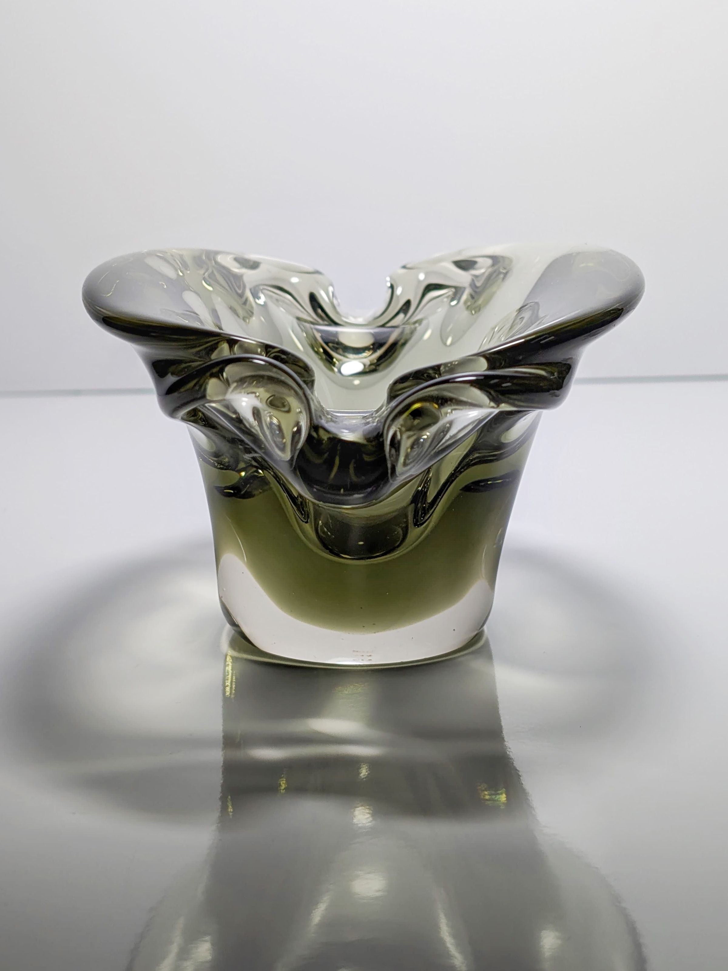 The Bohemian Smoked Glass Ashtray by Jan Beránek, available at Dodo Vintage, features a sculptural design with a fluid, wavy rim reminiscent of the 1960s. Its smoked glass and subtle gradients create an intricate play of light and shadow on glossy surfaces.