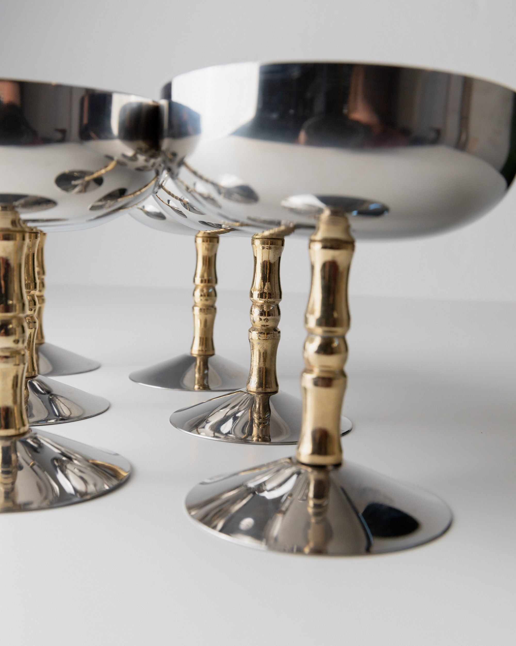 Close-up of Bottega Jacobs' 6 Stainless Steel Champagne Coupes 70s featuring metallic silver bowls and bamboo-like gold stems, arranged on a white surface. These vintage cups reflect light, showcasing their mid-century design elegance.