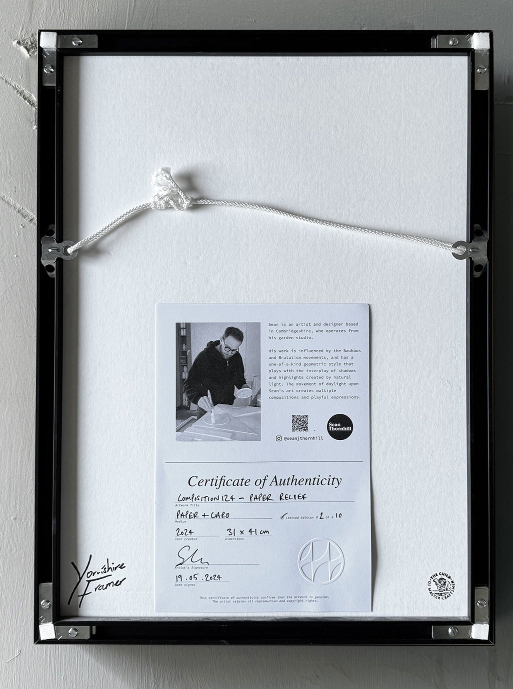 A framed certificate of authenticity labeled "Composition124" is secured with a white string on a gray wall. This limited edition piece from Sean Thornhill features a monochrome image of a person working on an art piece, accompanied by handwritten details and printed text.