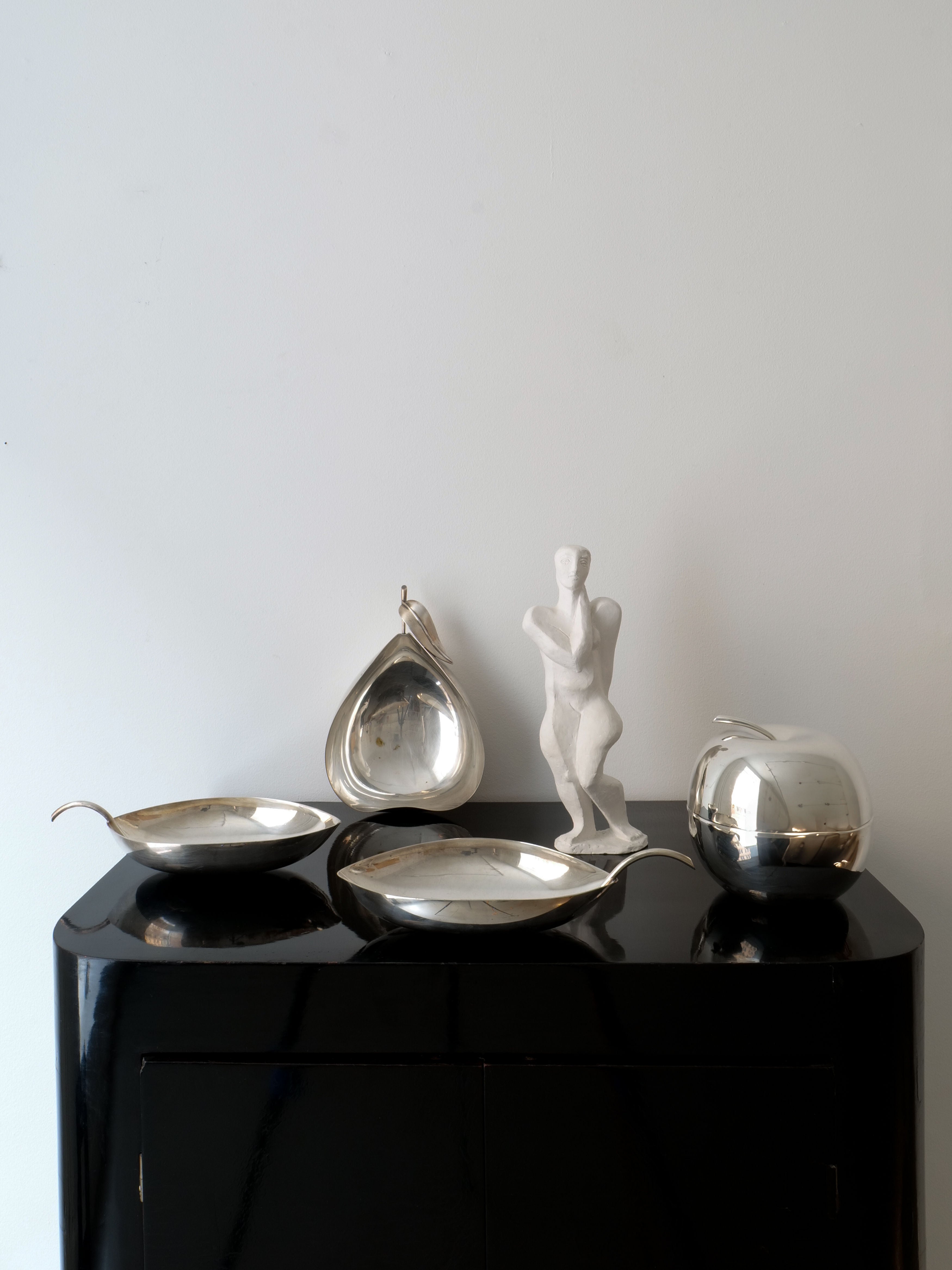 A minimalist display showcases Christofle Leaf Vide Poche bowls from Collection apart alongside a Lino Sabattini sculpture of a human figure, all set on a glossy black surface against a plain white background. This arrangement emphasizes a modern and artistic aesthetic.