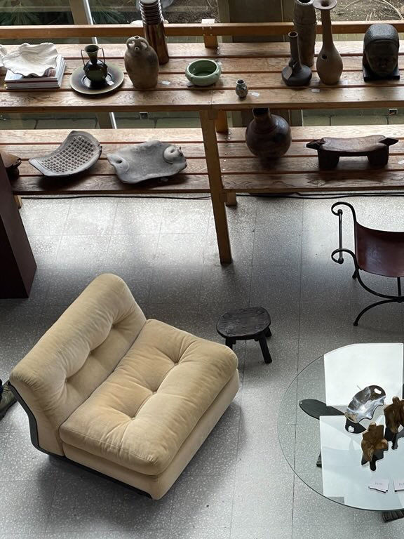 Two beige tufted Amanta armchairs designed by Mario Bellini in 1966 for RELIC LONDON sit on a gray tiled floor next to a small black wooden stool and a glass-top coffee table. Nearby, a wooden shelf showcases various ceramic and sculptural objects. A metal chair with a wooden seat is also visible in the stylish living space.