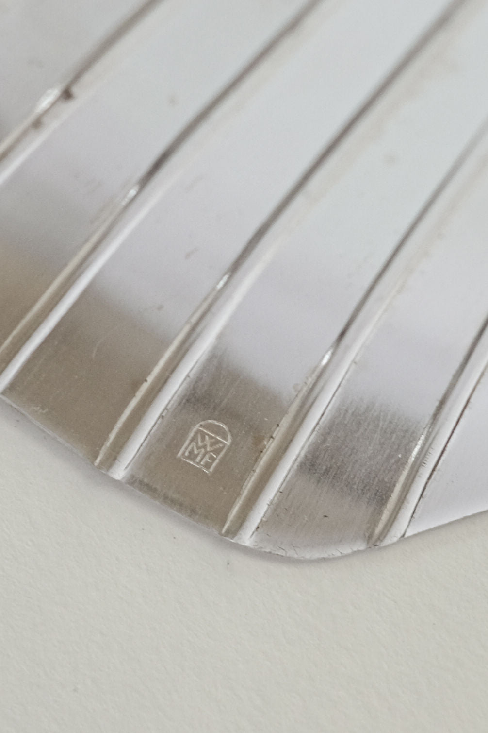Close-up of the Silverplated Shell Dish by septembre studios, featuring diagonal grooves for a vintage appeal. A stylized "WMF" logo is engraved near the edge in a trapezoid, contrasting elegantly against the plain background.