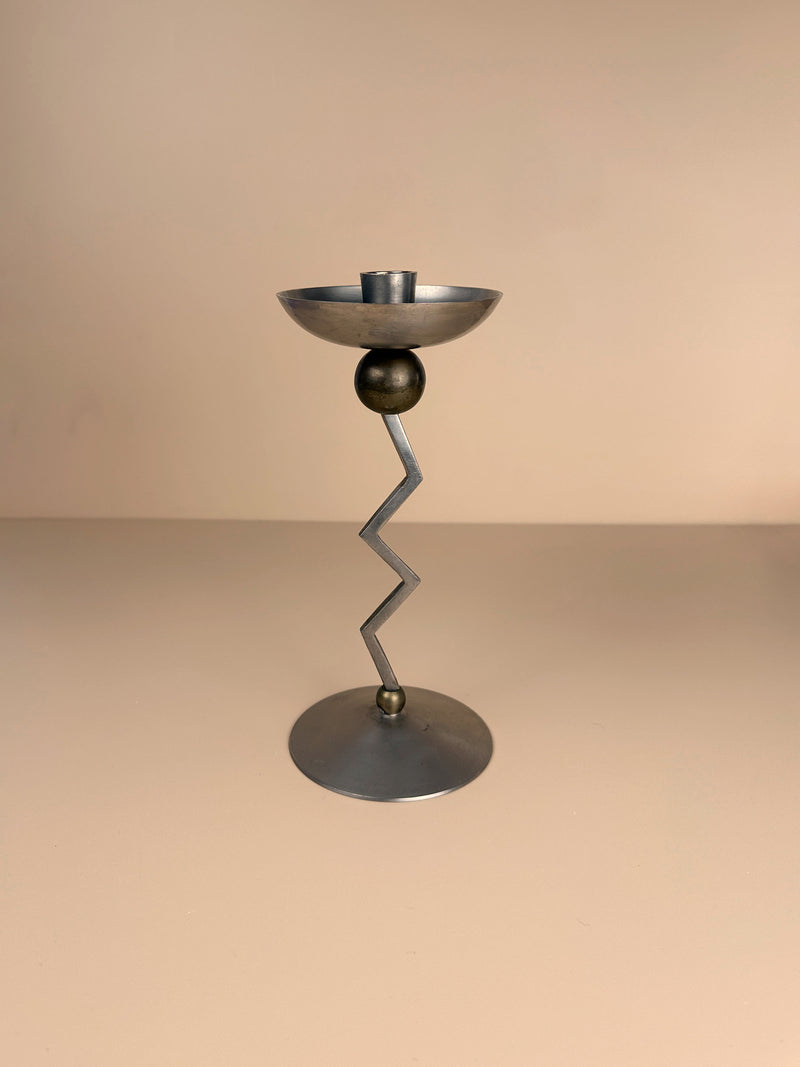 A contemporary, metallic candle holder with an elegant design, the Dodo Vintage Postmodern Zig Zag Candle Holder features a round base, a zigzag-shaped stem, and a shallow circular holder at the top. It is expertly crafted from silver-colored metal against a solid, neutral background.