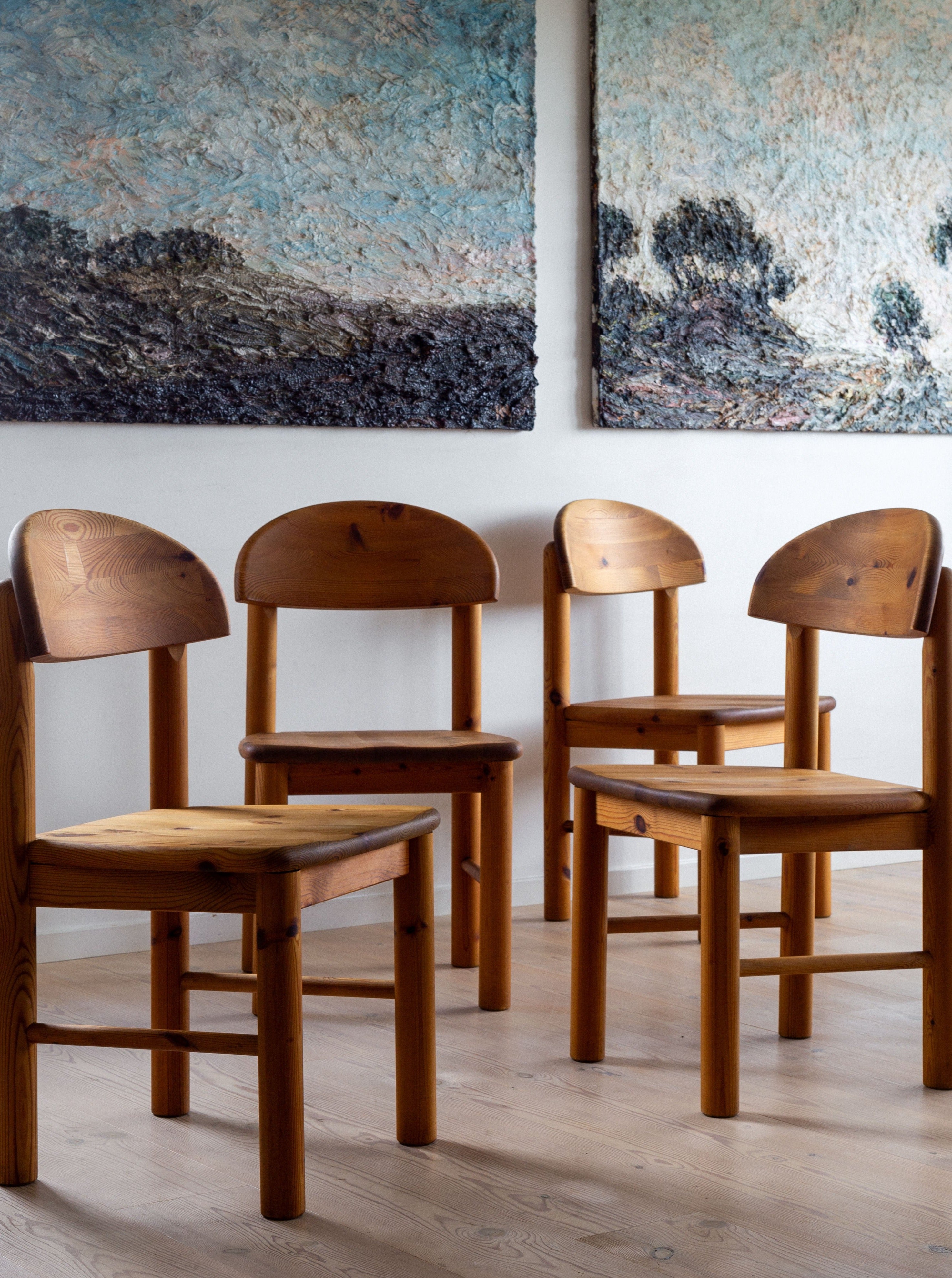 Four Pine Chairs by Rainer Daumiller from the 1970s, crafted in Denmark and branded by Spigel, with their rounded backs and seats are arranged on a light wood floor. Two large textured landscape paintings, featuring trees and a cloudy sky, hang on the wall in the background. The scene is well-lit, highlighting the natural wood grain of the solid pine furniture.