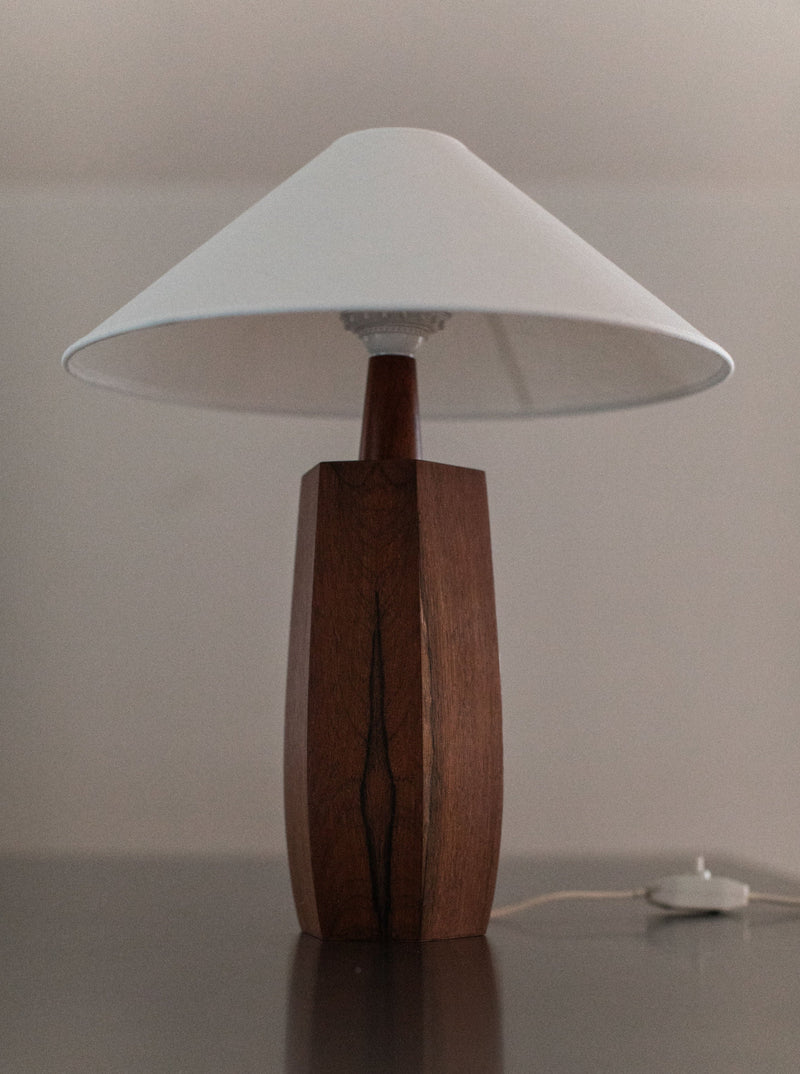 The 1940's Wooden lamp by Out For Lunch exemplifies mid-century design with its wooden, hexagonal base, complemented by a white conical lampshade. Positioned on a reflective dark surface, its meticulous craftsmanship is evident. A white power cord is visible at the base against a plain, light-colored background.