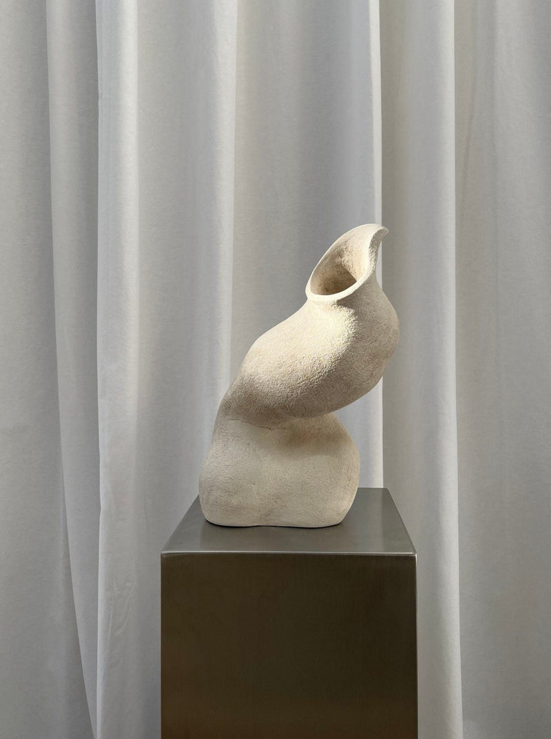 An abstract Sculpture Seashell by 2222Studio, resembling a curved, swooping form resting on a metallic pedestal against a backdrop of soft, flowing white curtains.