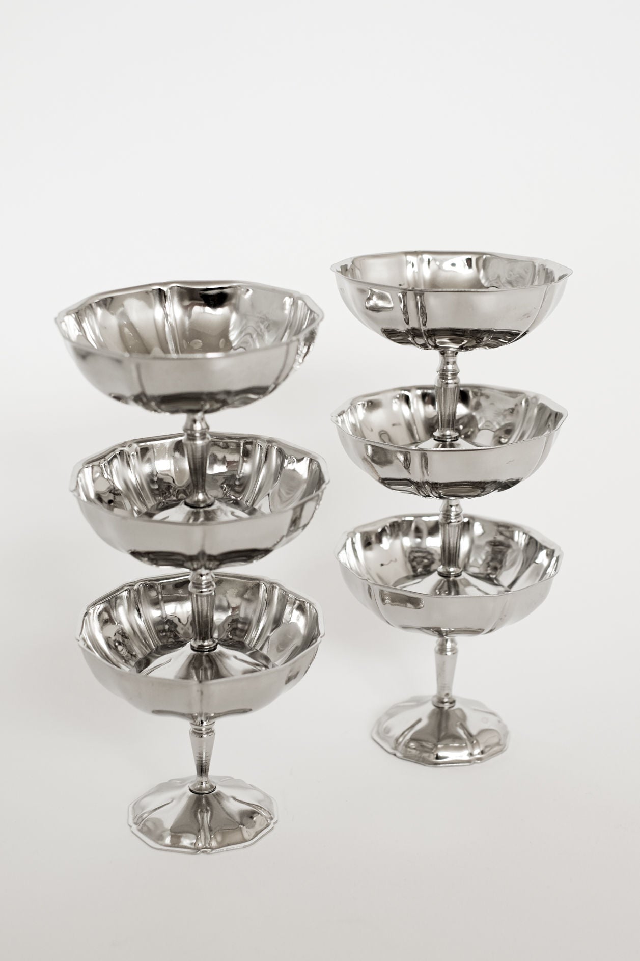 Six shiny stainless steel coupe glasses from septembre studios are arranged in two columns on a plain white background, with each stack featuring two glasses balanced for vintage appeal.