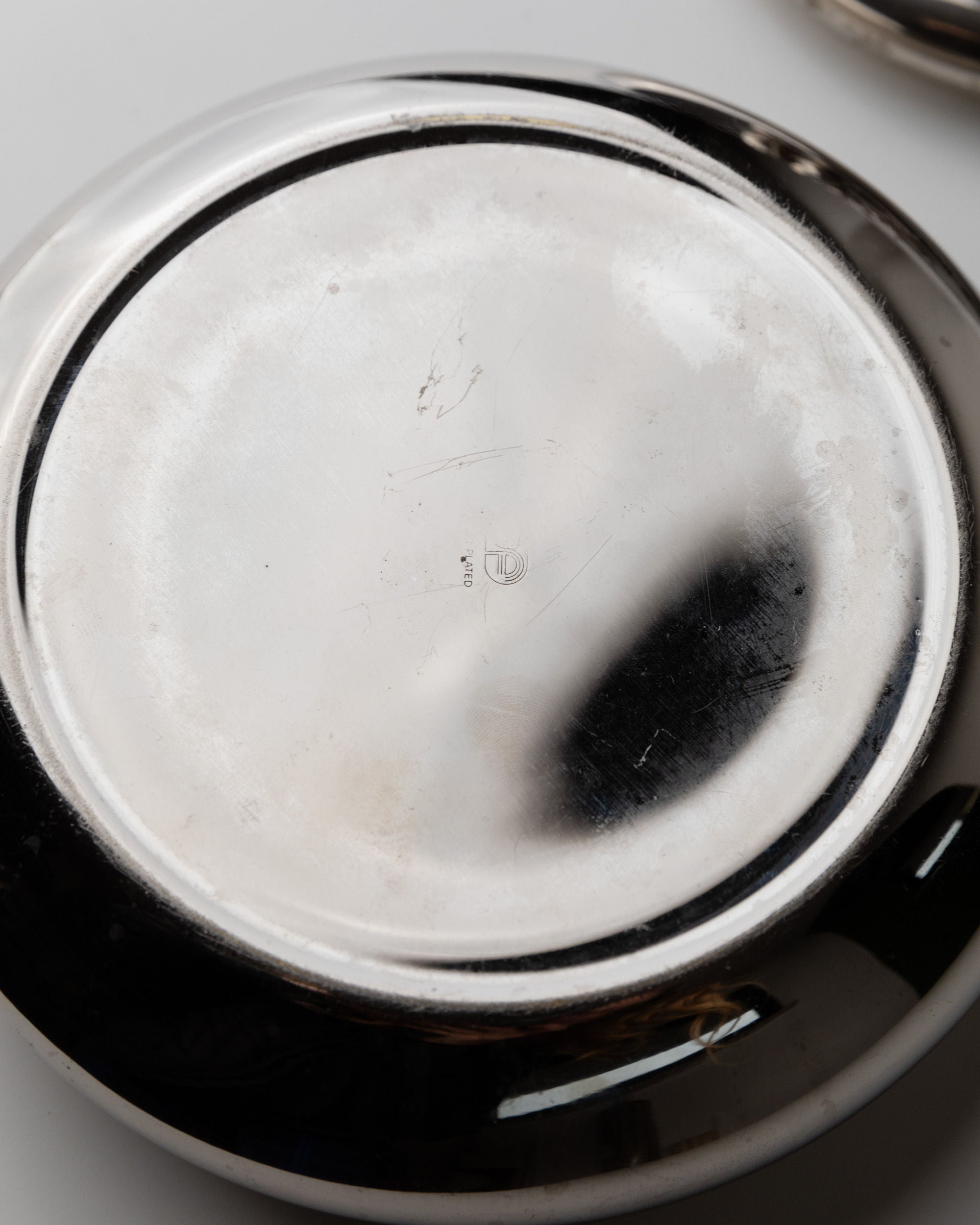 The image shows the underside of a circular, silver-plated metallic object from Bottega Jacobs. The Silver-plated Box from Italy's 1950s collection features a reflective surface with scratches, a dark smudge on one side, and a faint marking at the center against a plain background.
