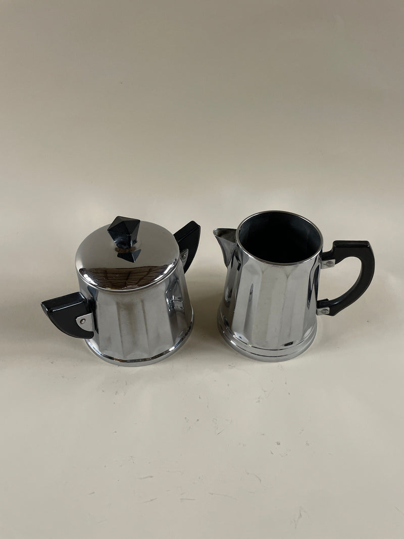 Two sleek, silver Art Deco Sugar Bowl and Jug by Maud Vaughan grace a neutral background, exuding a vintage French art deco flair. The sugar bowl on the left is topped with a lid, while the jug on the right remains open, both boasting a reflective surface akin to chrome.