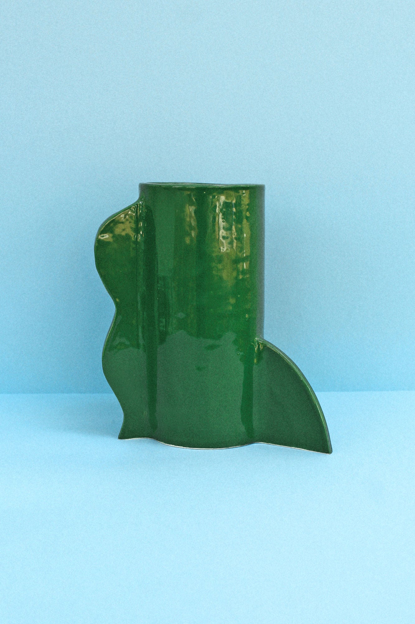 A green ceramic Theatre Vase 04 (Bosque) by Casa Veronica with an artistic, abstract shape sits on a light blue surface against a light blue background. Hand-built earthenware, the vase has a glossy finish and features unique, irregular curves and edges.