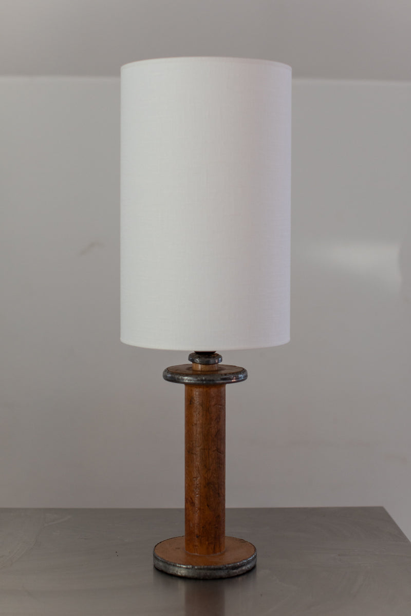 An Out For Lunch Large Mid-Century Lamp featuring a white cylindrical shade and a wooden handle base sits on a dark, reflective surface. The plain and neutral background enhances the minimalist and modern aesthetic of the overall setting.